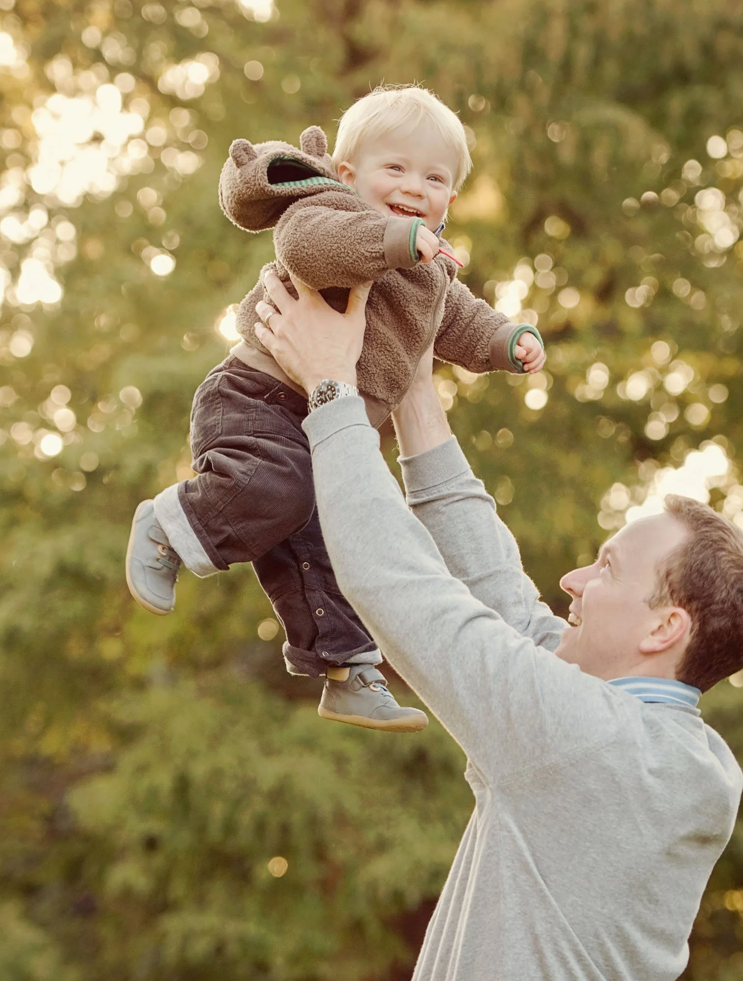 A 1 year old boy laughs as he's held up in the air by his smiling proud father.  The golden sunlight shines through the leaves behind them.