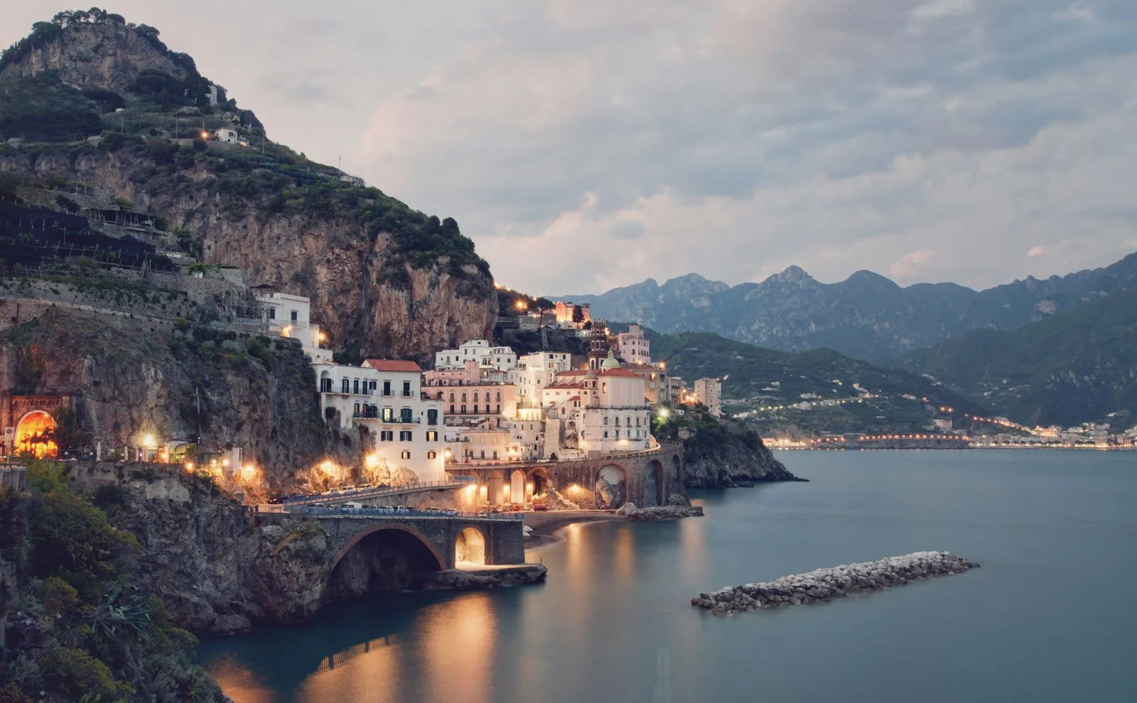 Coastal town at dusk with buildings on cliffs, a curved bridge, calm water, and mountains in the background.