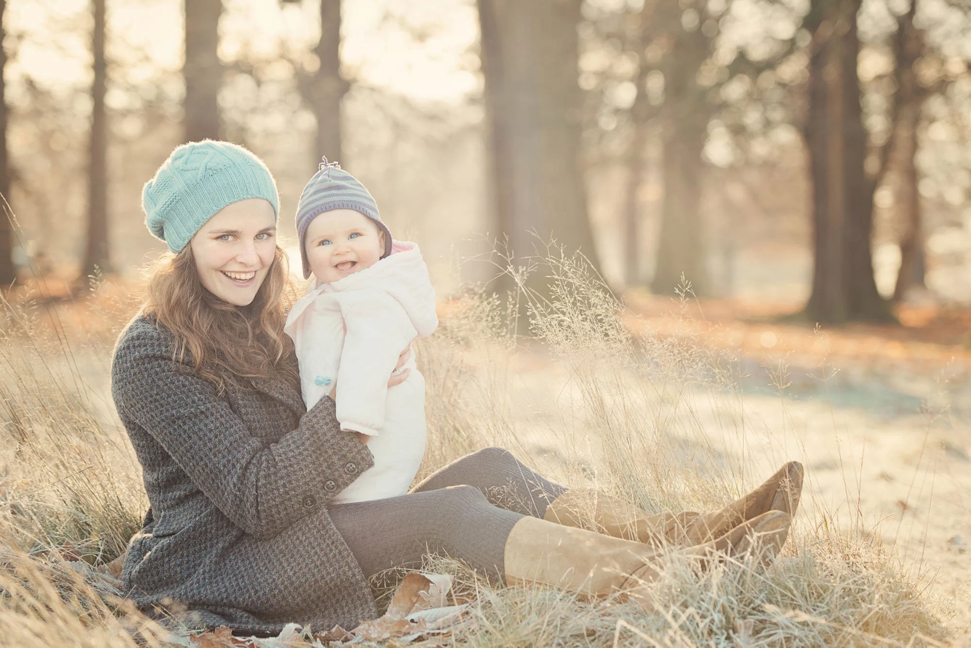 A proud mother sits on the ground and holds her baby as both smile at the camera.  Both are warmly dressed for the winter photoshoot in Hampstead Heath as the winter sun shines through the trees behind them.