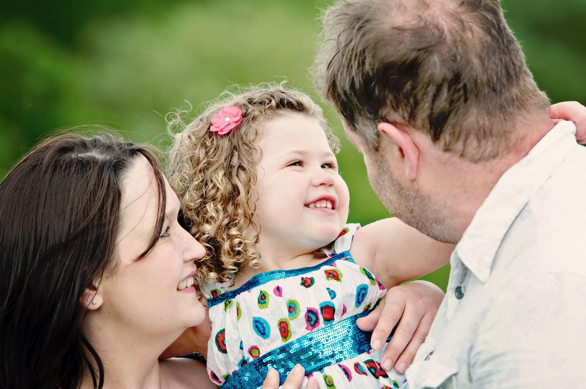 Little girl is hugged by her parents and smiles at her dad during a family photoshoot in Regent's Park London