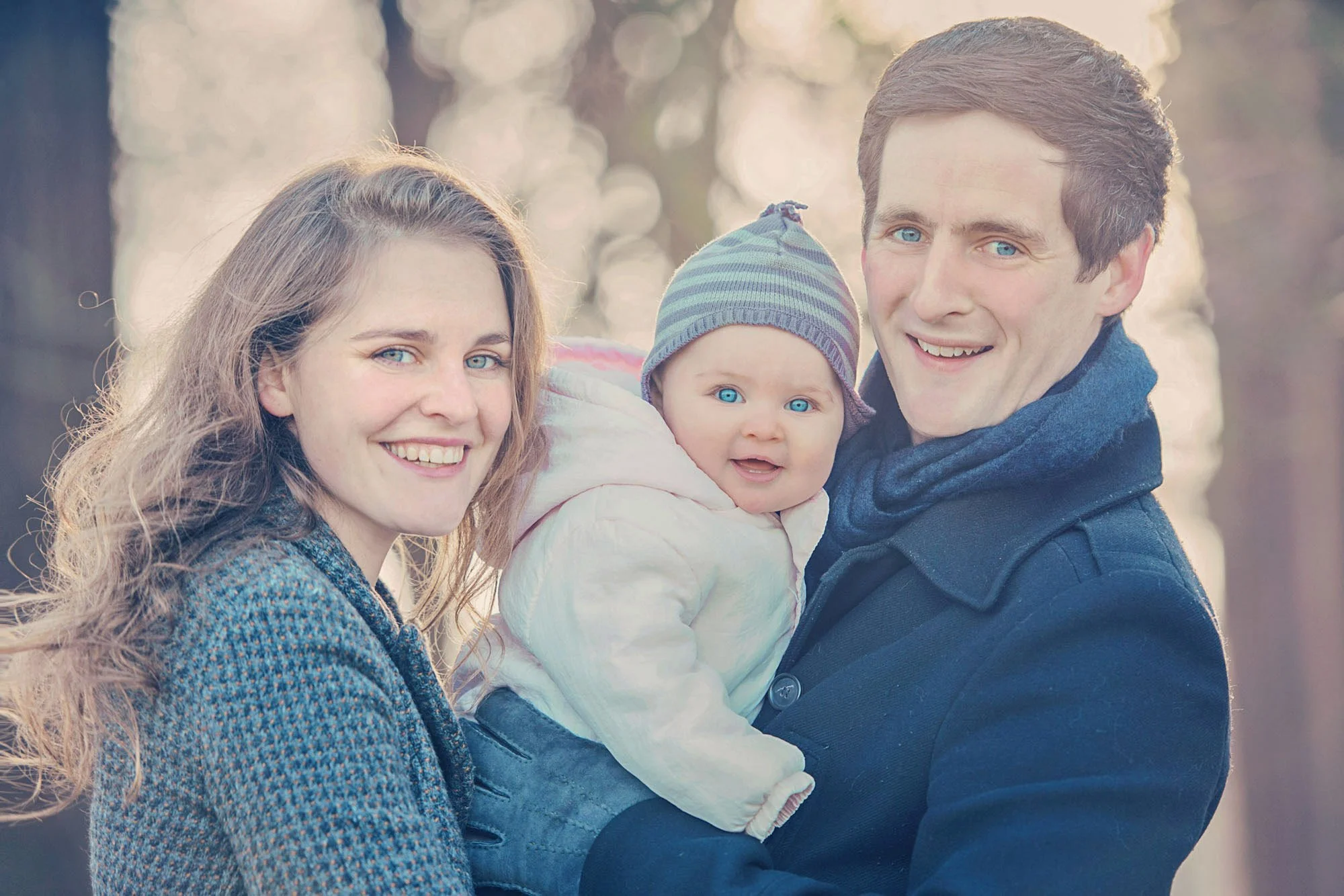 A baby with blue eyes and her parents smile at the photographer during a winter family photoshoot in Richmond Park.  The parents are wearing winter coats and the baby wears a hat.