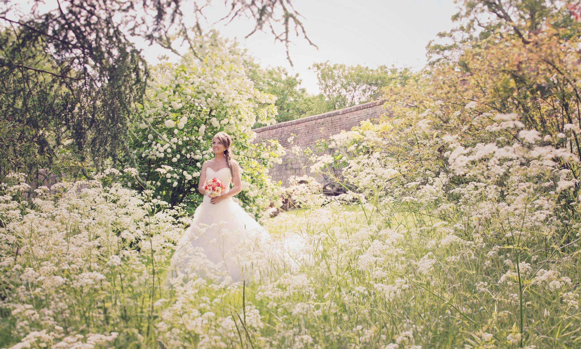 A bride stands in the garden among a profusion of white flowers during her weekday wedding at Farnham Castle in Surrey