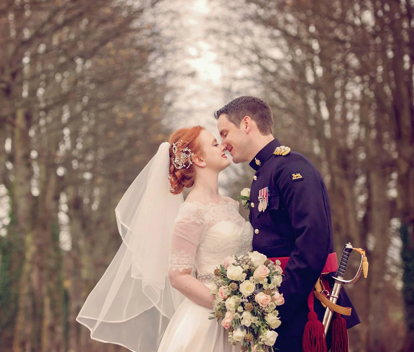 A groom in full uniform kisses his red-headed bride as she holds her bouquet and they stand under an avenue of bare trees during the winter in St James' Park in London