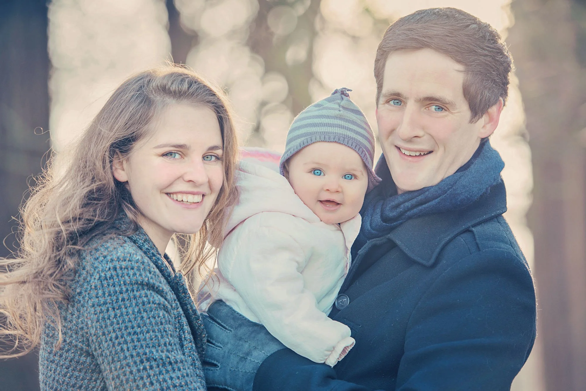A six month old baby and her parents smile at the camera during a winter photoshoot in Richmond Park in west London.  The baby wears a hat. 