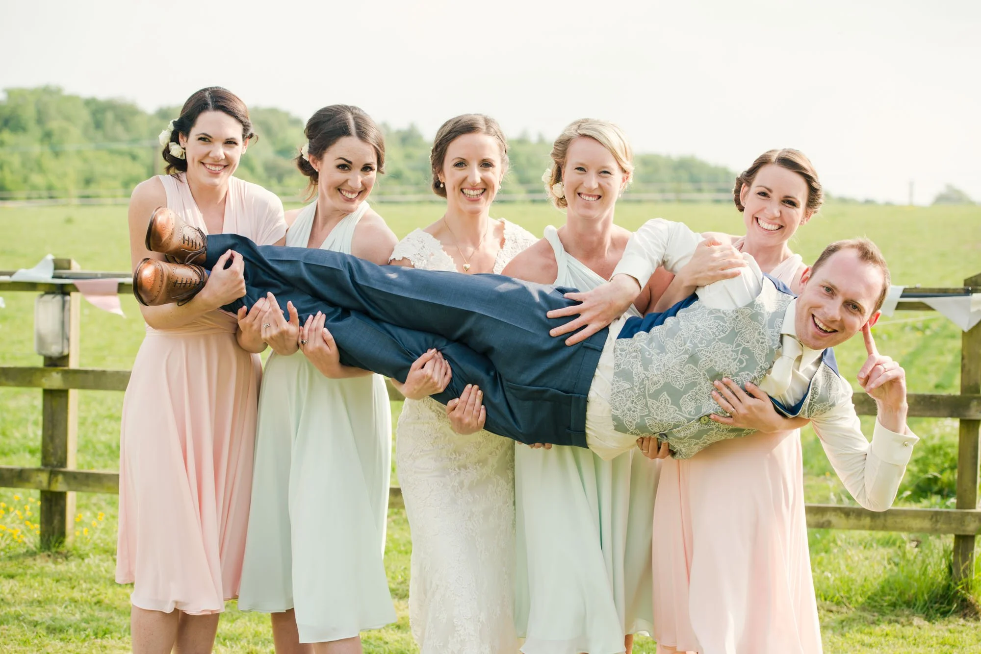 A playful group of bridesmaids holds up the groom for a group photo.  They are standing in front of a fence and field during a wedding reception held at a farm in Kent  