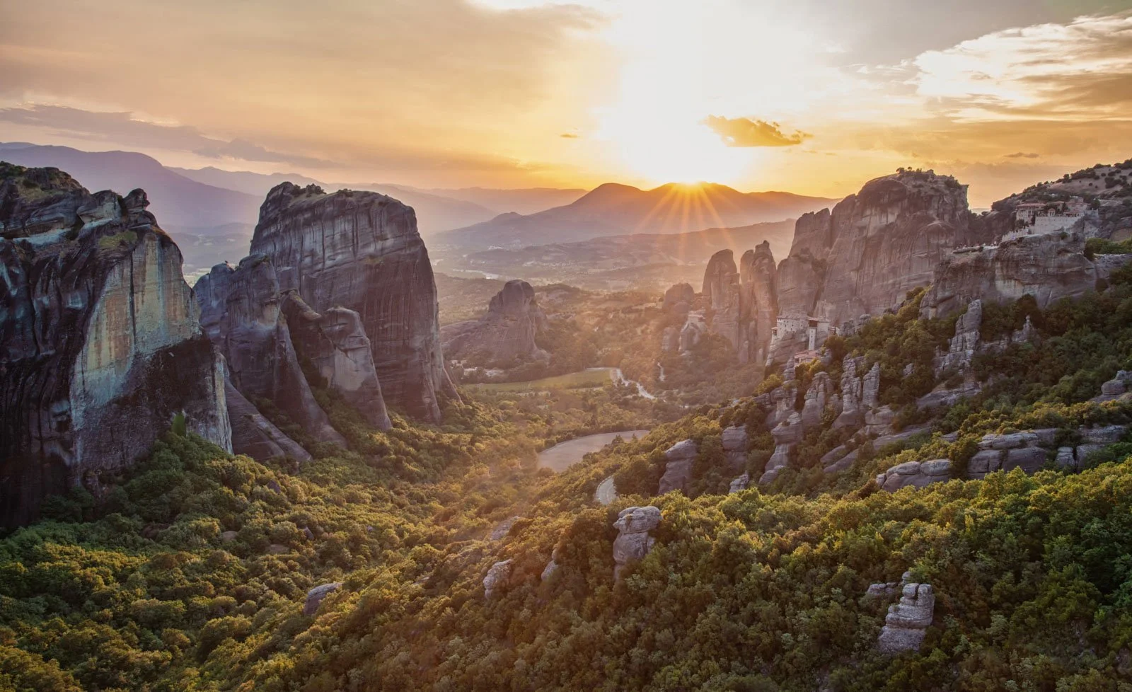 A mountain landscape at sunset with large rocky formations, lush green forest, and a winding road or river in the valley.