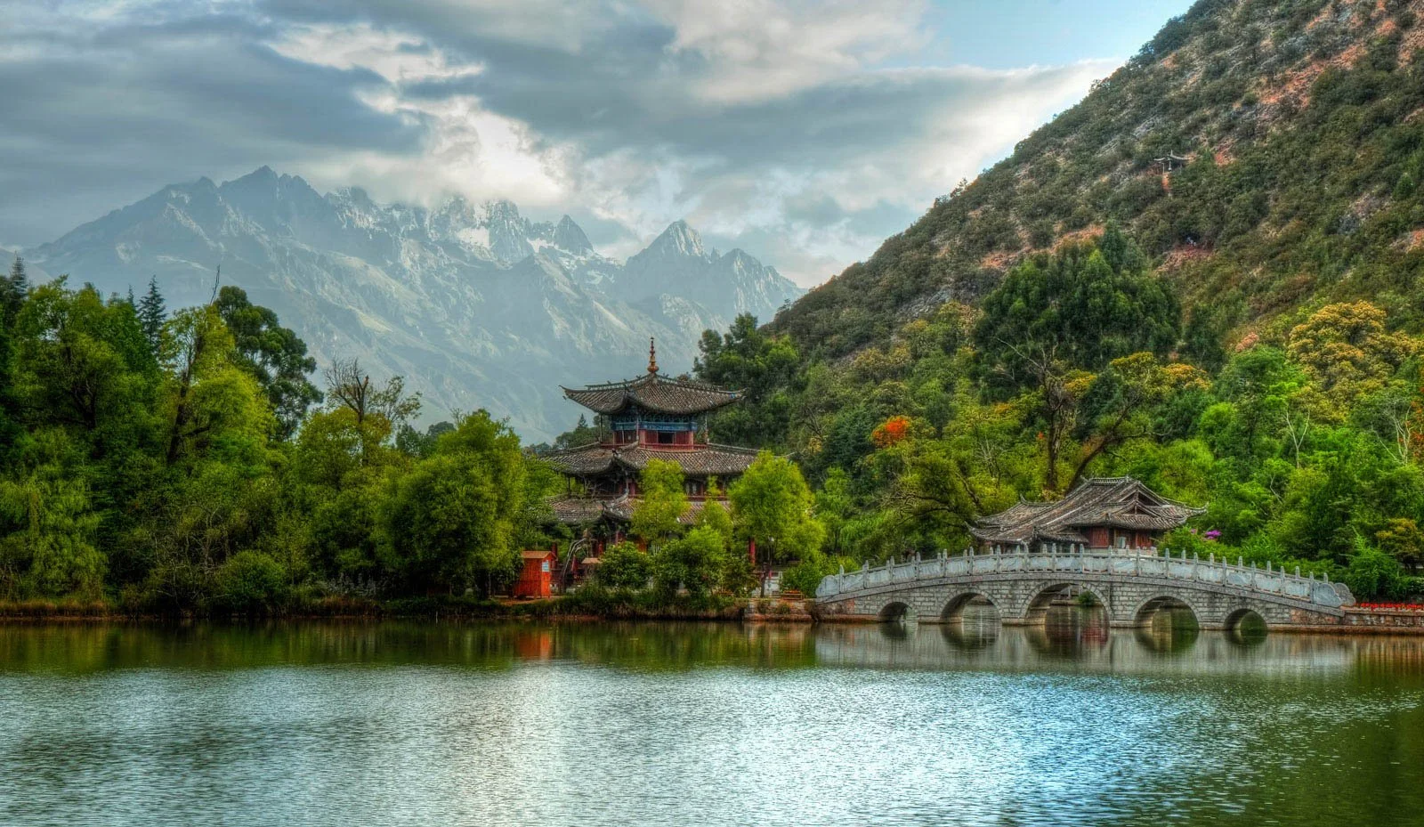 Traditional Asian pagoda-style building located on the bank of a lake, with a stone bridge arching over the water and lush green trees, mountains in the background, and a partly cloudy sky overhead.