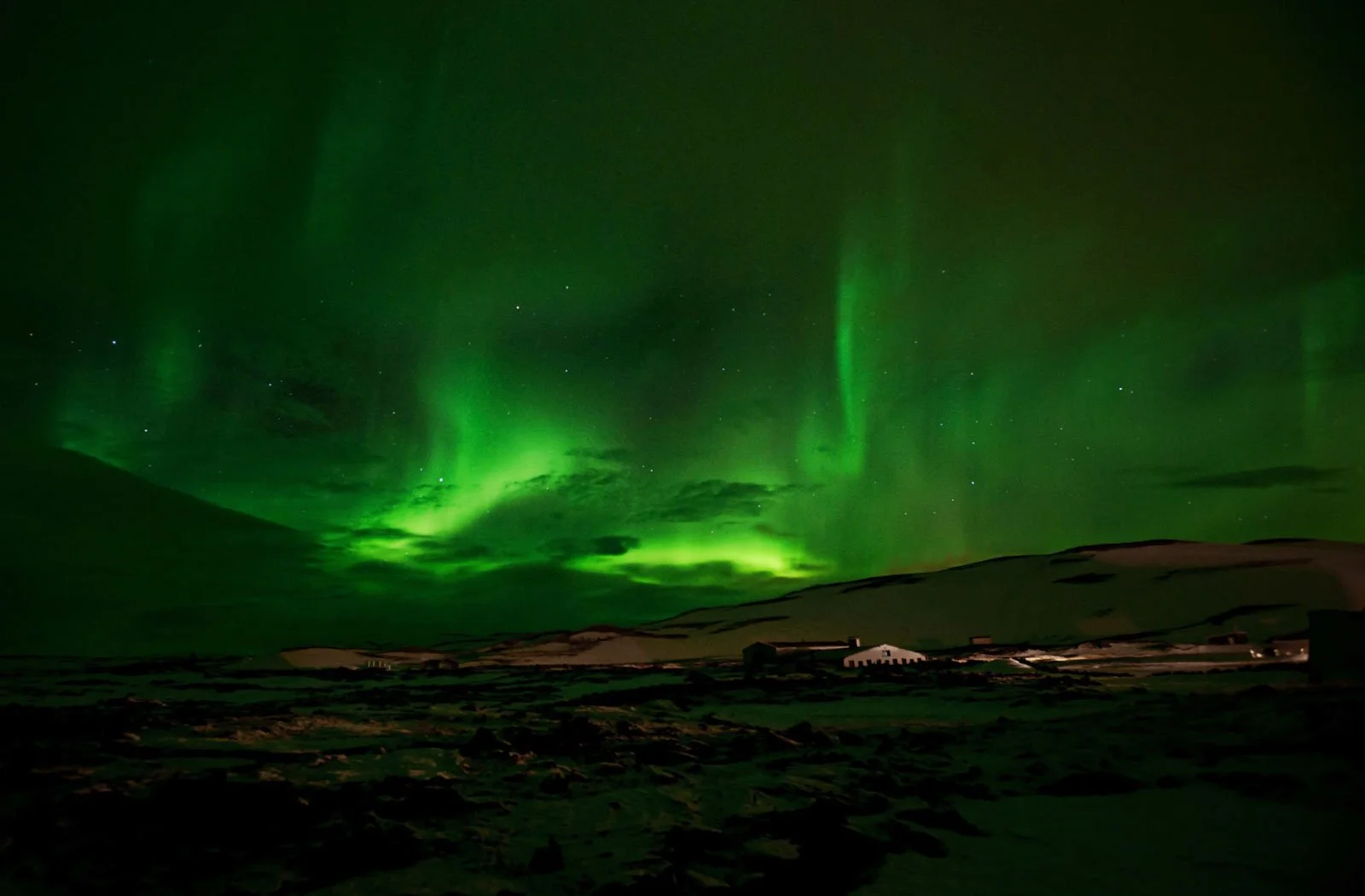 Northern lights glow green over a snow-covered landscape at night with some buildings in the distance.