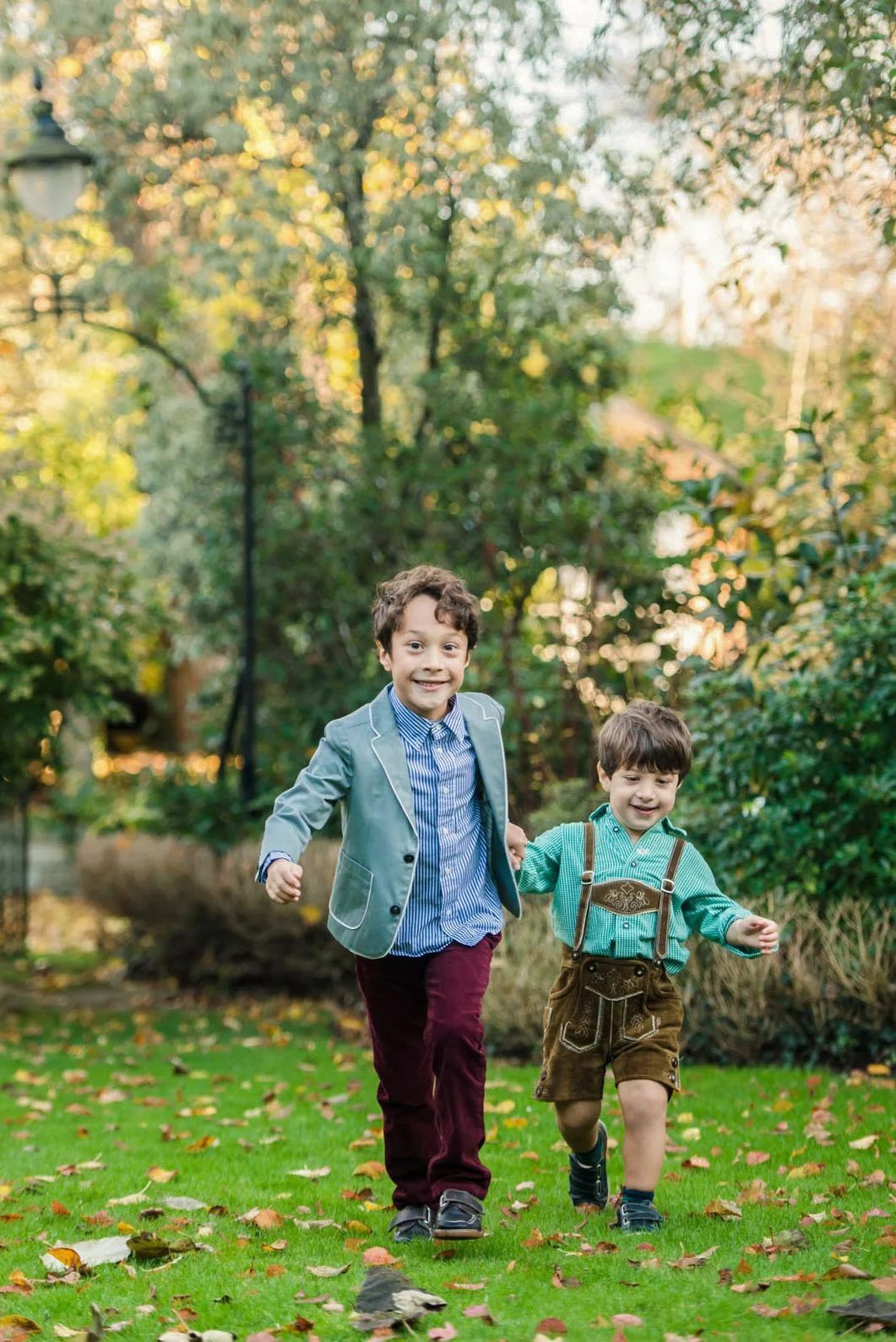 A little boy, smartly dressed in shirt and jacket, holds his little brother by the hand and leads him across the grass towards the photographer. This family photoshoot was held in the garden of a family home in Hampstead.