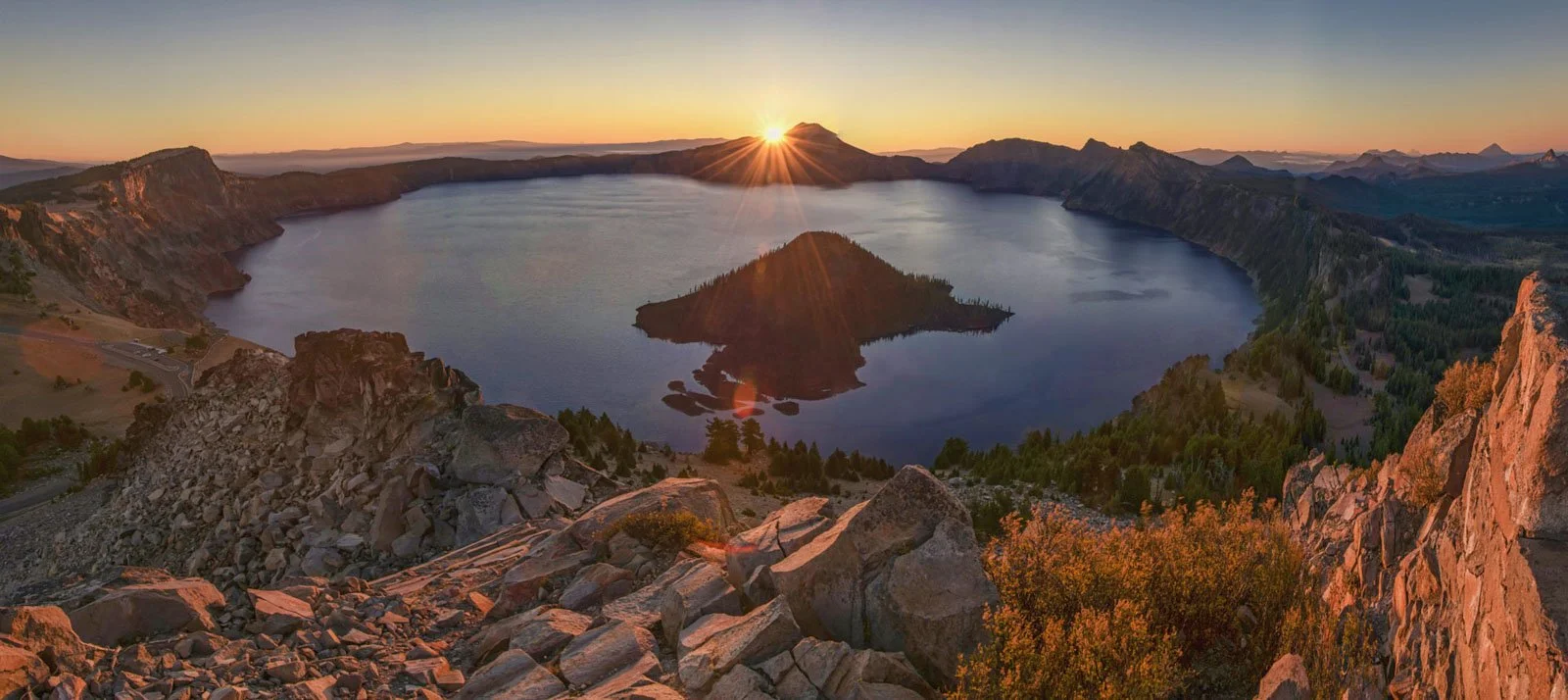 Sunset over Crater Lake with a volcanic island in the center, surrounded by cliffs and forested slopes.