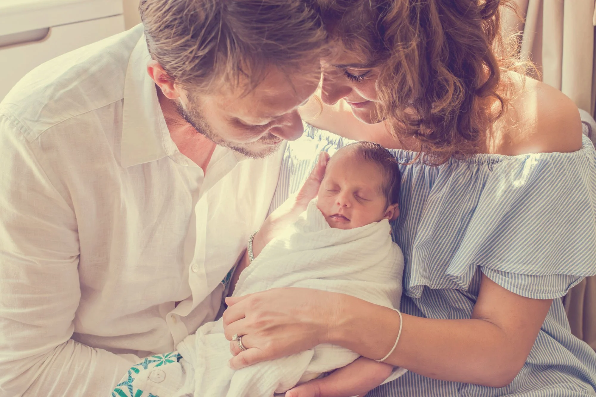 A sleeping newborn baby lies peacefully asleep as she is held by her parents in a moment of love and peace.  Warm sunlight bathes the family.
