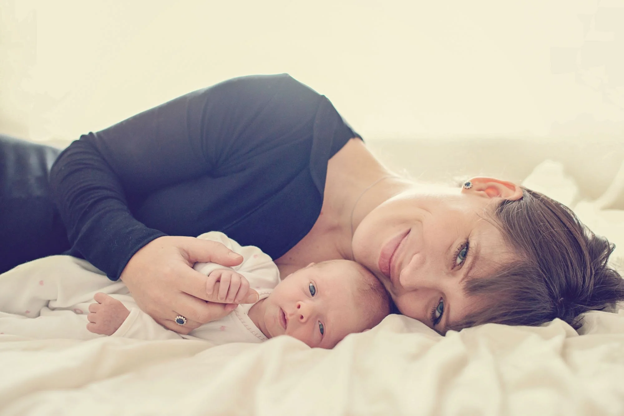 A proud and serene new mother lies on her side with her newborn baby, and holds her baby's hand.  Both mother and baby look at the camera, during a newborn shoot in Hampstead in north London.