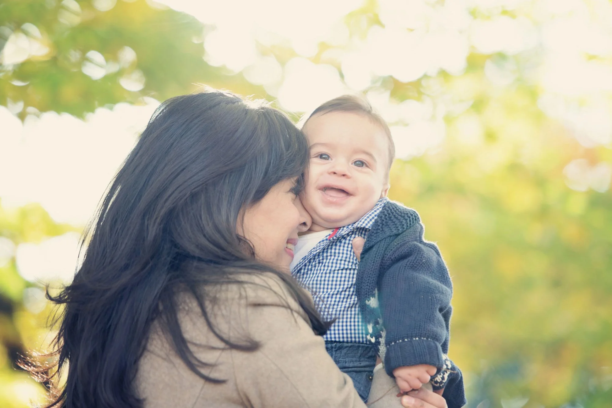 Proud mother cuddles her baby son as he laughs in delight, during a family photoshoot in Hyde Park in London, with dappled light shining through the yellow foliage behind them. 