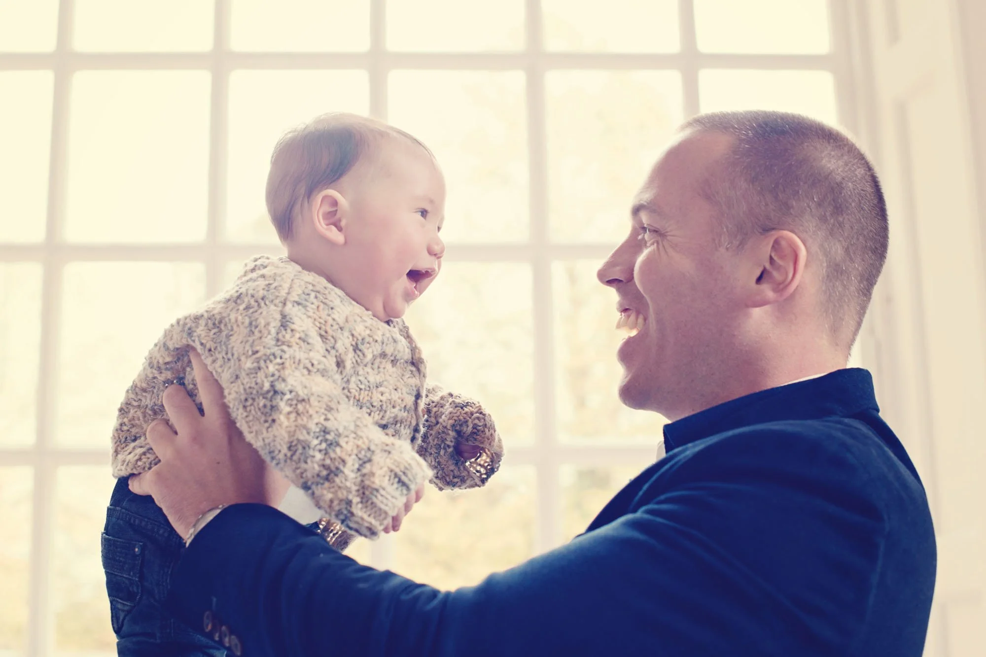 A proud father holds his six month old baby boy up in front of him and they smile delightedly at each other in the front room of their family home in Notting Hill in London