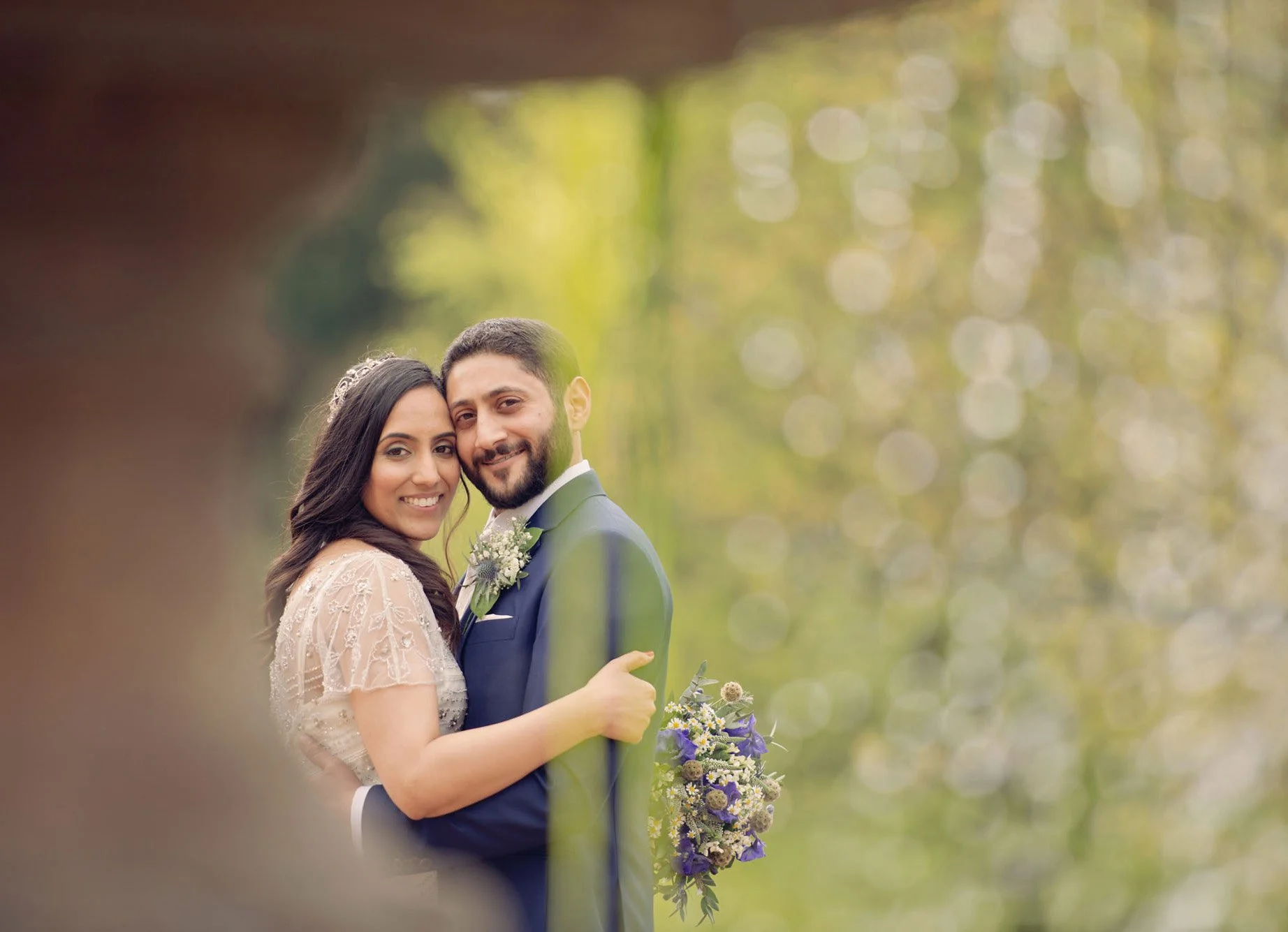 A bride and groom hold each other and smile at the camera as they stand behind a fountain in Regent's Park after their micro wedding at Camden Register Office