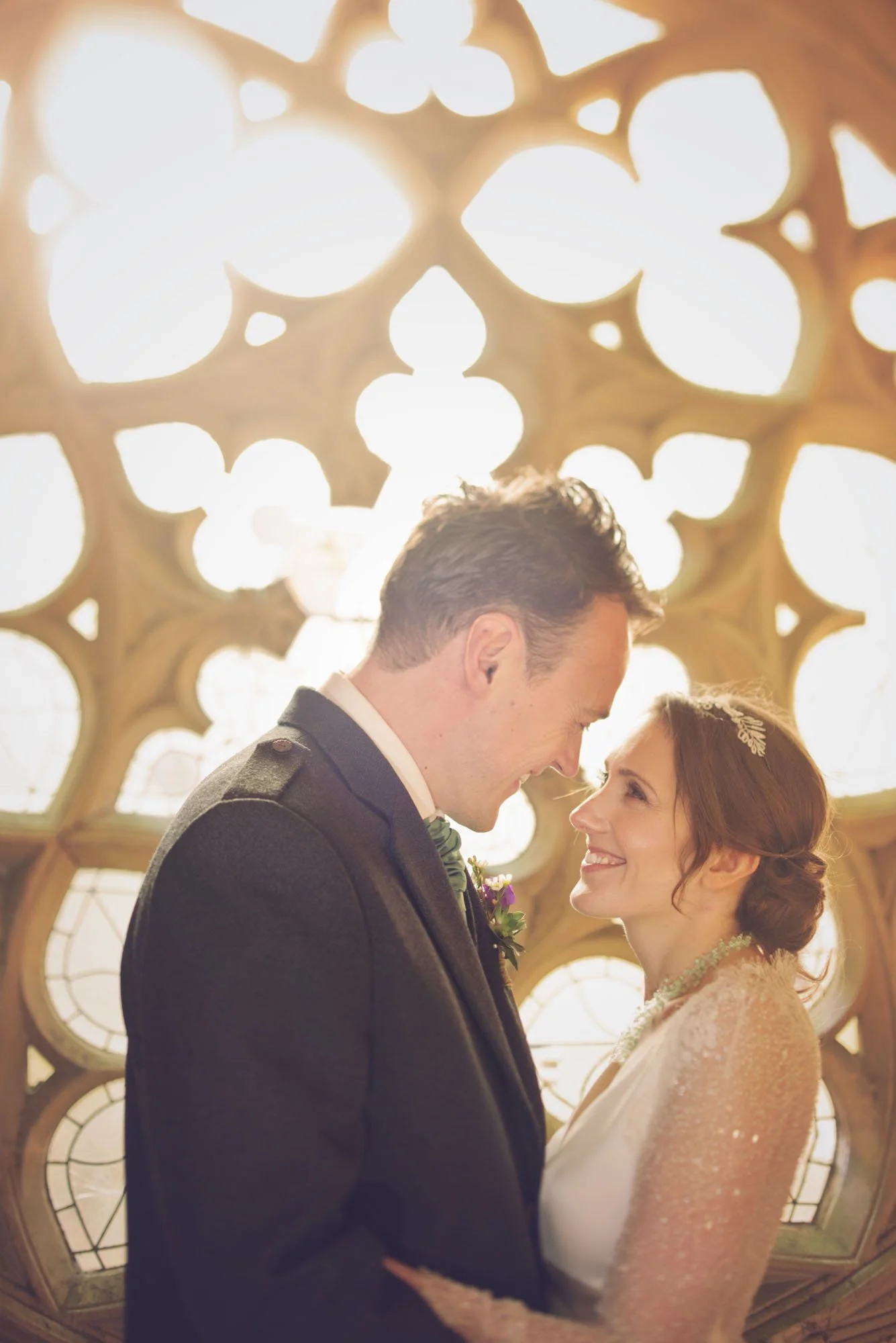Bride and groom smile at each other with happiness as they stand in front of a giant window in London with ornate patterning.