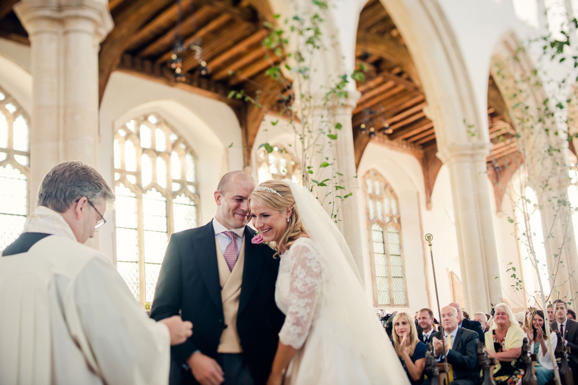 Bride and groom lean into one another smiling as they stand before the priest in a bright and airy London church in which saplings have been placed down the aisle.