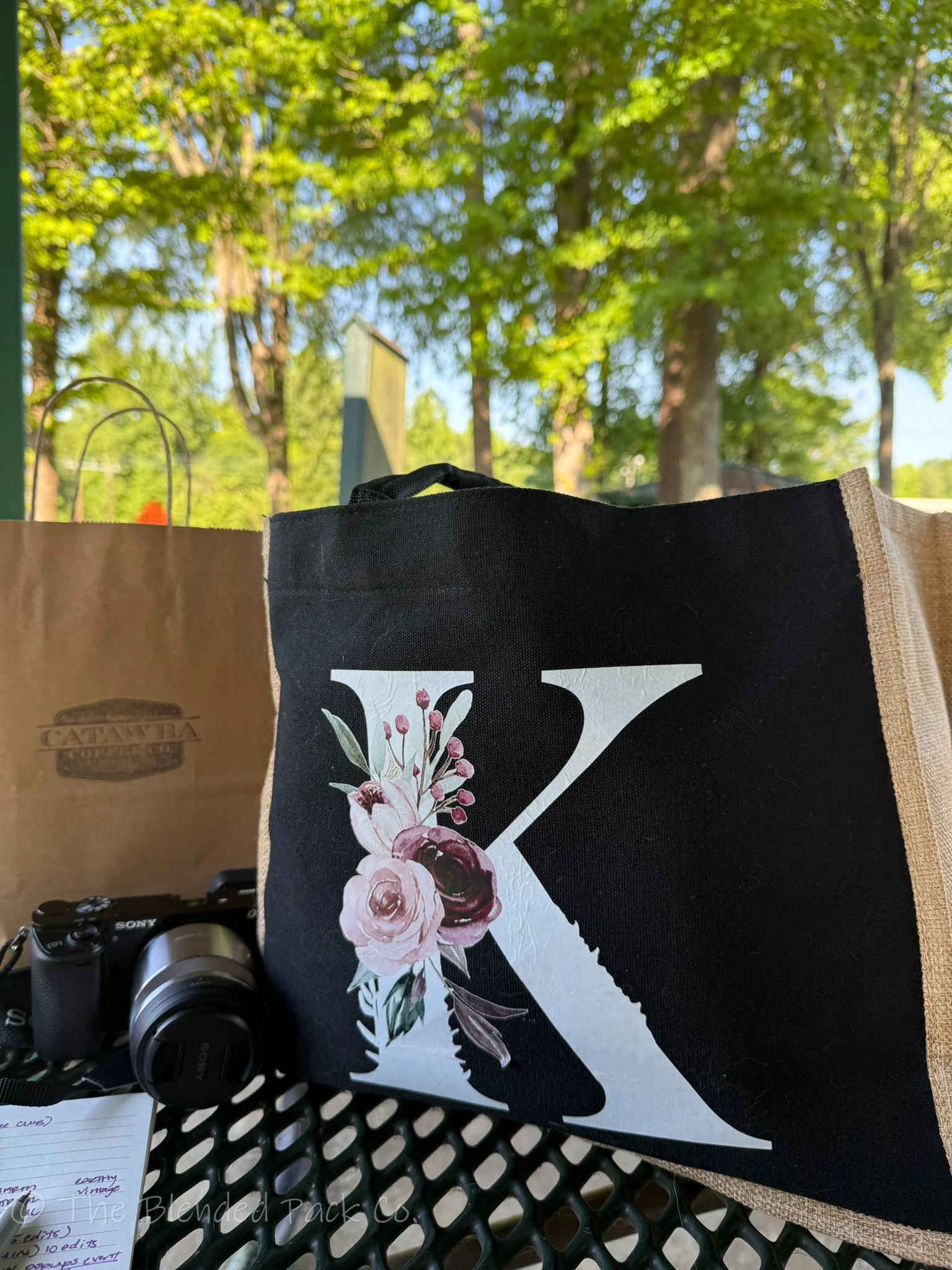 A black tote bag with a floral design and a white silhouette of a chainsaw, placed on a black metal outdoor table. In the background, there are green trees and a blue sky. A camera is partially visible at the bottom left corner of the image.
