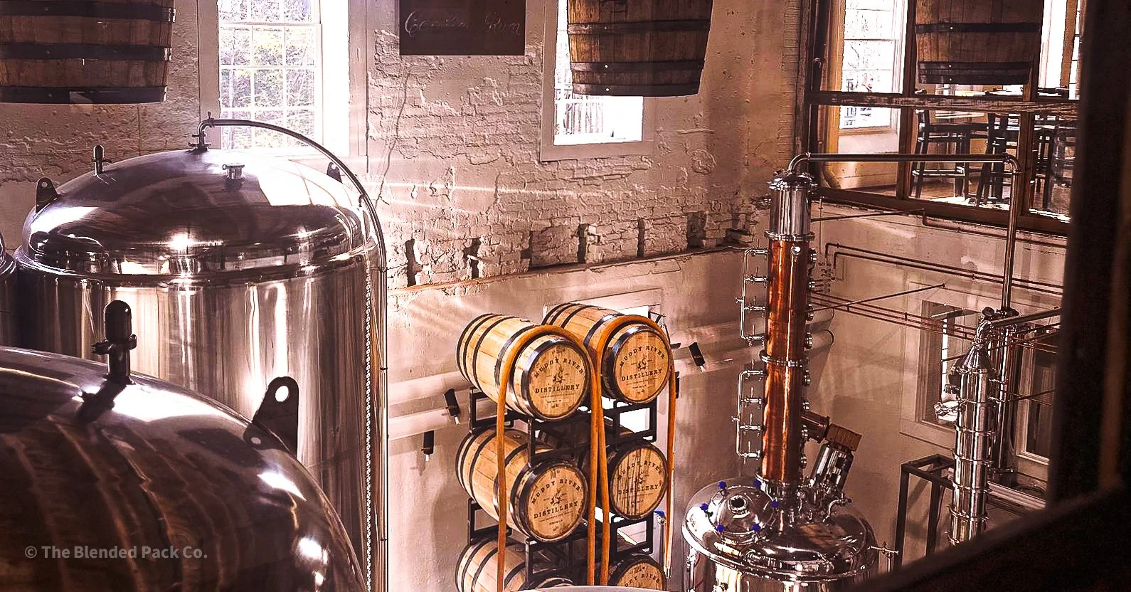 Inside a distillery with large metallic tanks, shelves of wooden barrels labeled 'Happy River Distillery,' and copper pipes against a brick wall.