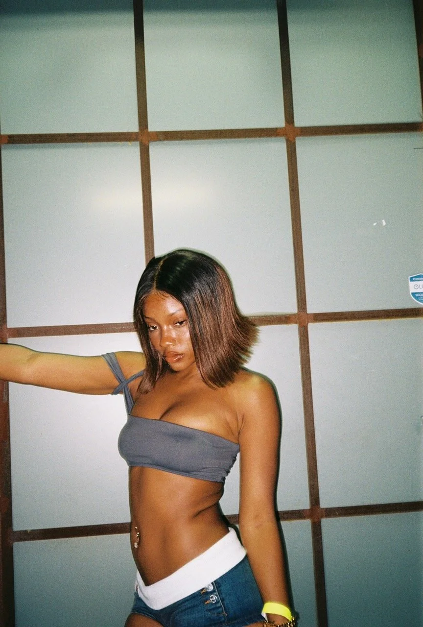 Young woman with shoulder-length hair, wearing a gray crop top and blue shorts, standing in front of a wooden wall, posing with one arm extended outward.