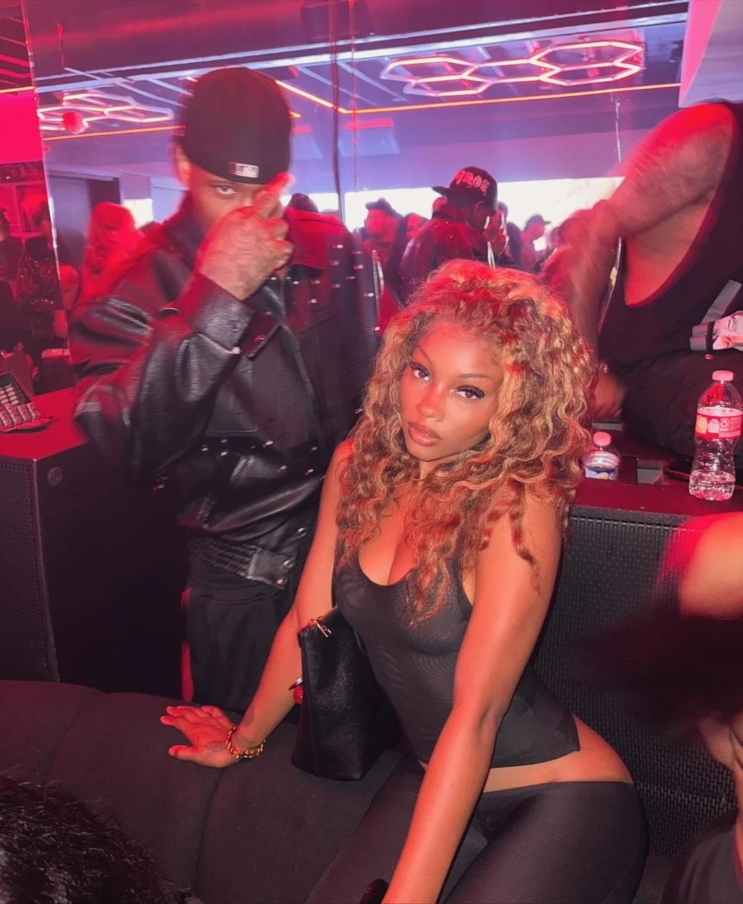 A woman with curly blonde hair sits on a black couch in a nightclub, looking at the camera with a pose. A man in a black leather jacket and black cap stands nearby, partially flipping the camera the middle finger. The background shows a crowded space with red and blue neon lighting and other people socializing.