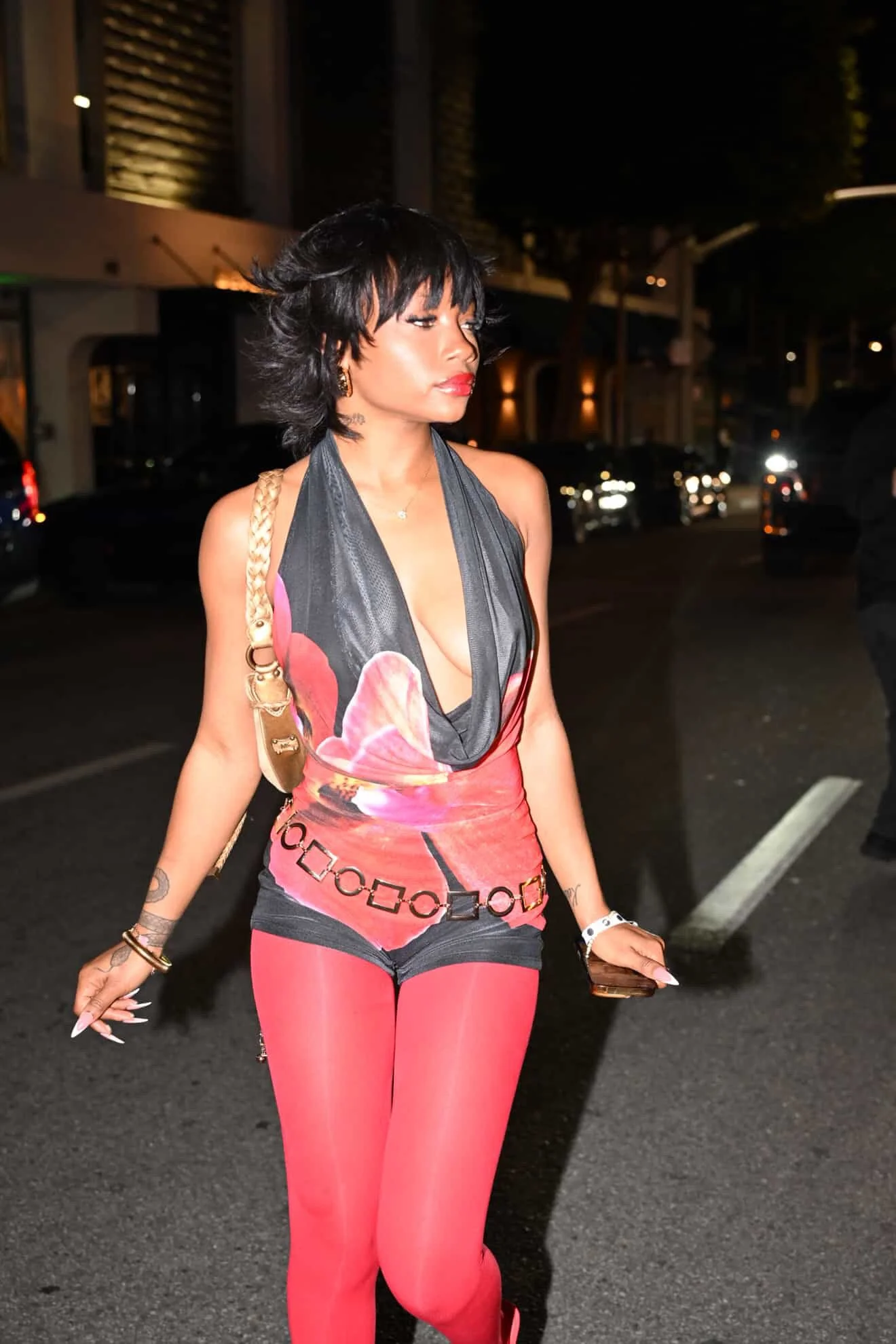 A woman walking on a city street at night, wearing a colorful dress and pink leggings, holding a smartphone, with blurred city lights in the background.