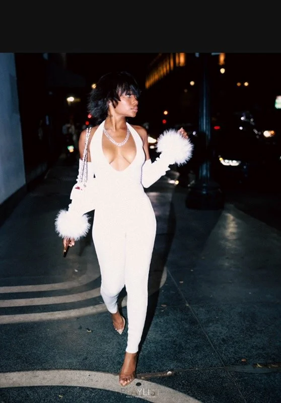 Woman in a white jumpsuit with long white gloves and fluffy cuffs, walking on a city sidewalk at night.