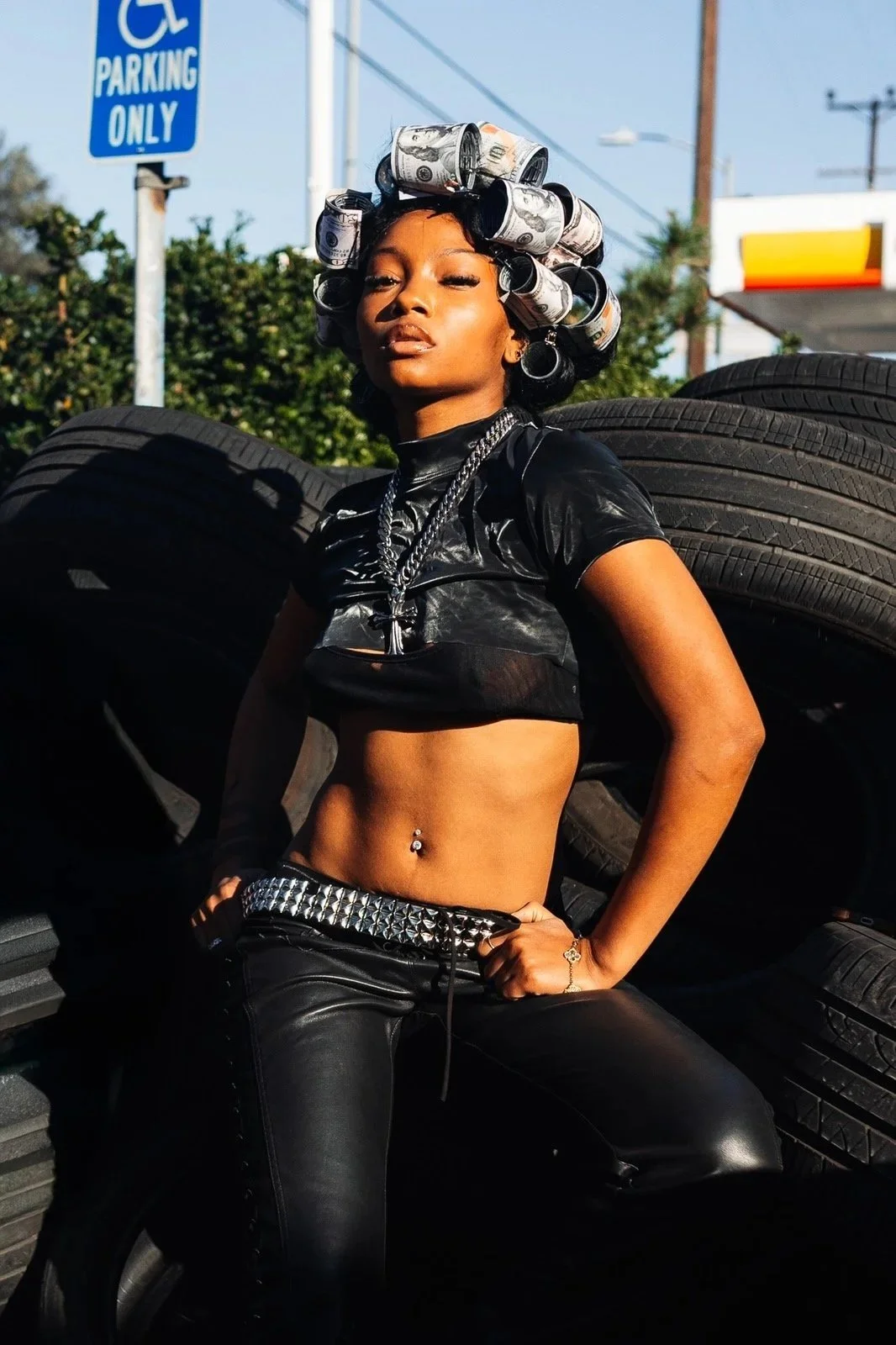 A woman with curled hair made of rolled dollar bills, wearing a black crop top and leather pants with a studded belt, standing in front of stacked tires outdoors.