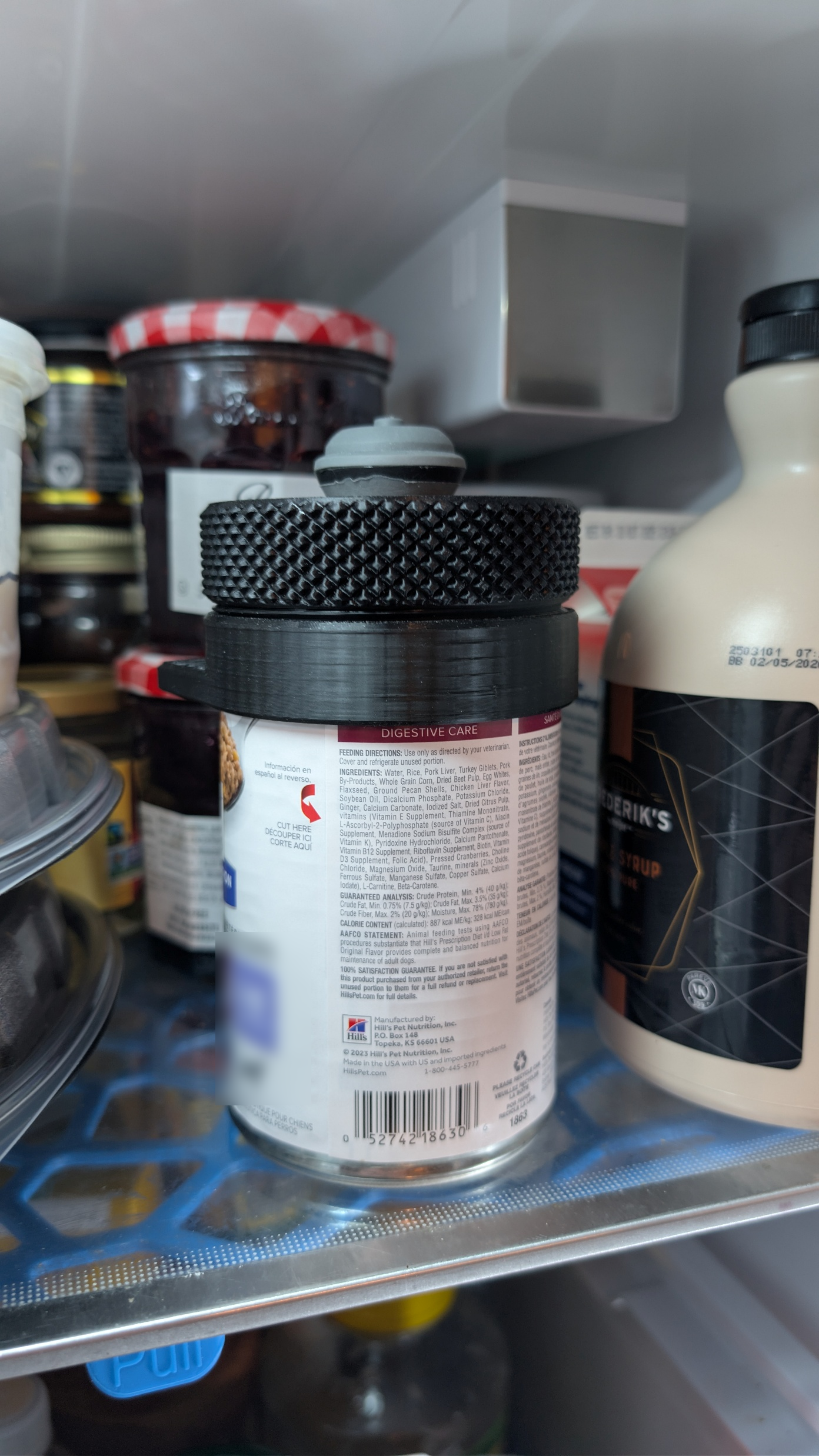A pet food can with a black textured lid in a refrigerator, surrounded by other jars and bottles.