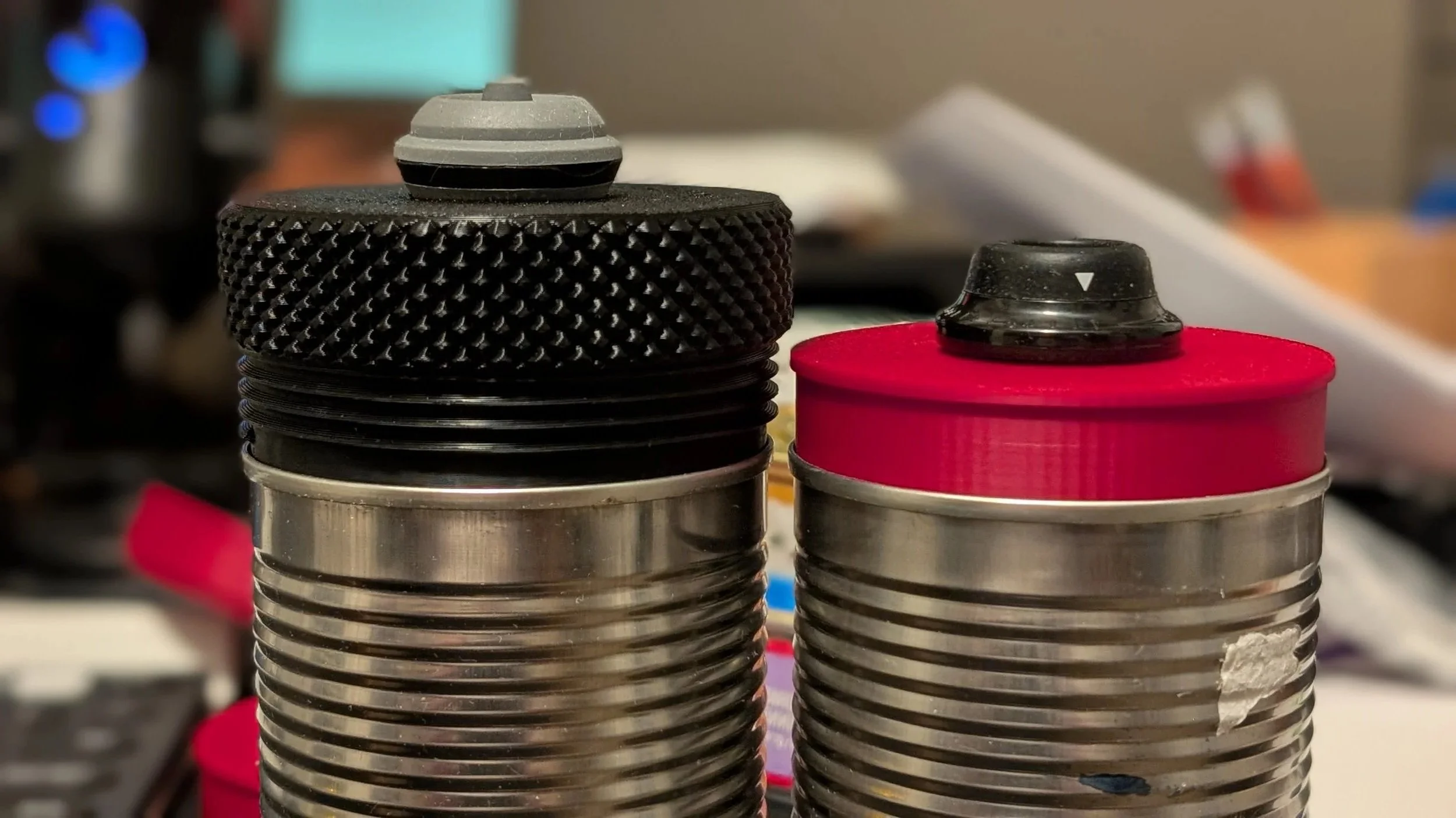 Close-up of two metal cans fitted with prototype lids and vacuum pump valves on top, one can with a gray valve and the other with a black valve, in a workshop or kitchen setting.