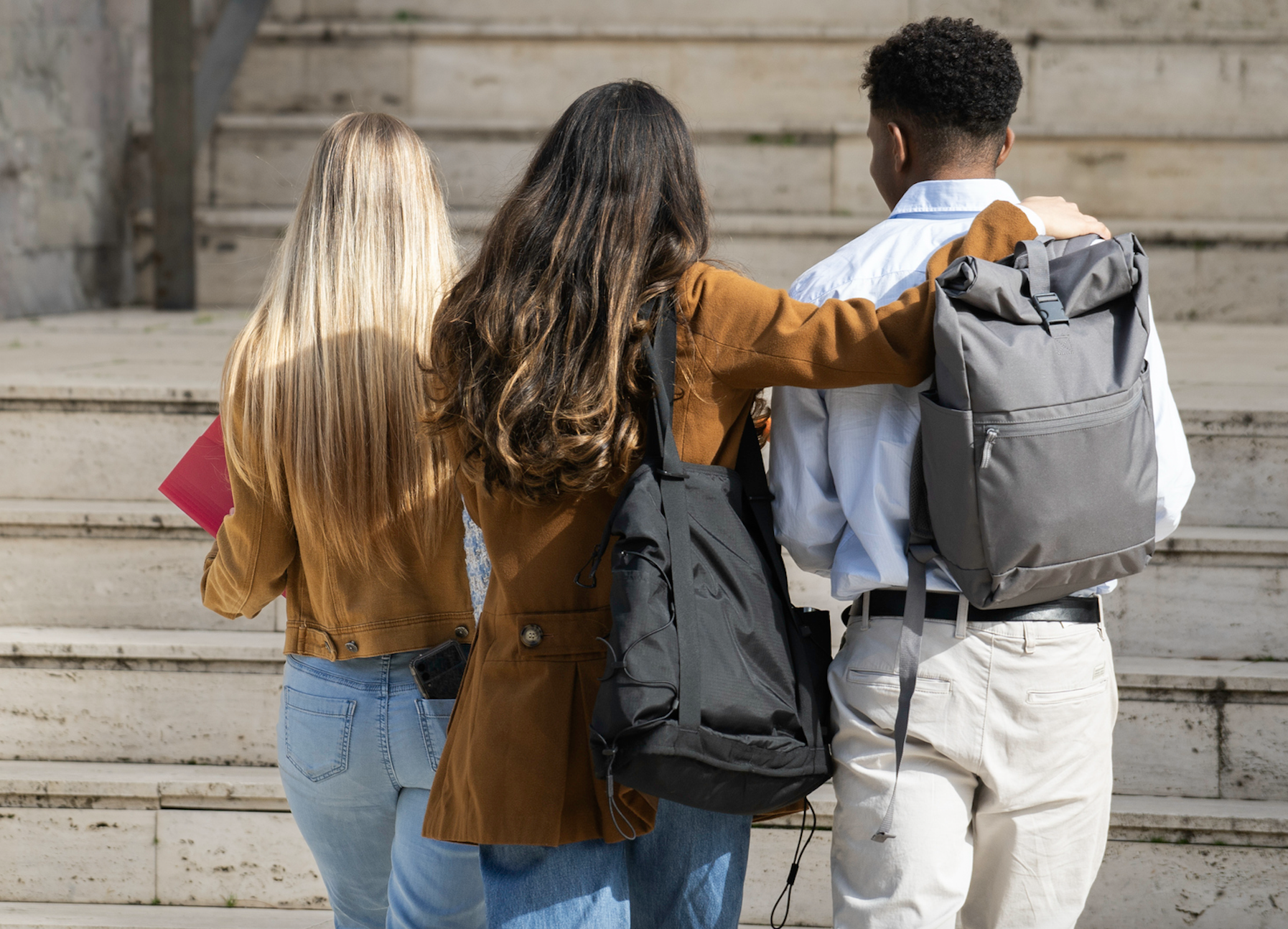 Three teenagers, two girls and one boy, walking up outdoor steps with their arms around each other's shoulders, carrying backpacks and a folder.