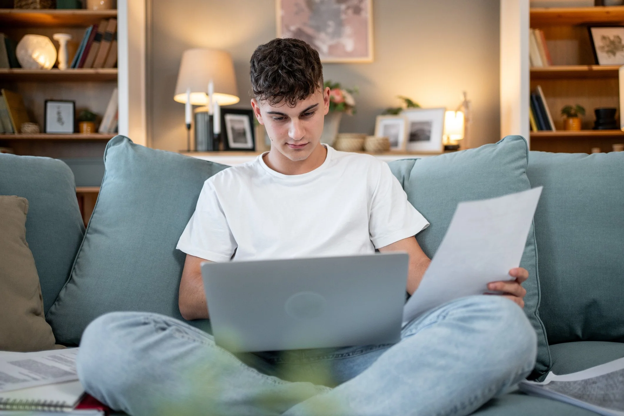 A young man with curly hair wearing a white t-shirt sitting on a gray sofa, looking at papers in his hand in a cozy living room with bookshelves, lamps, and framed photos in the background.