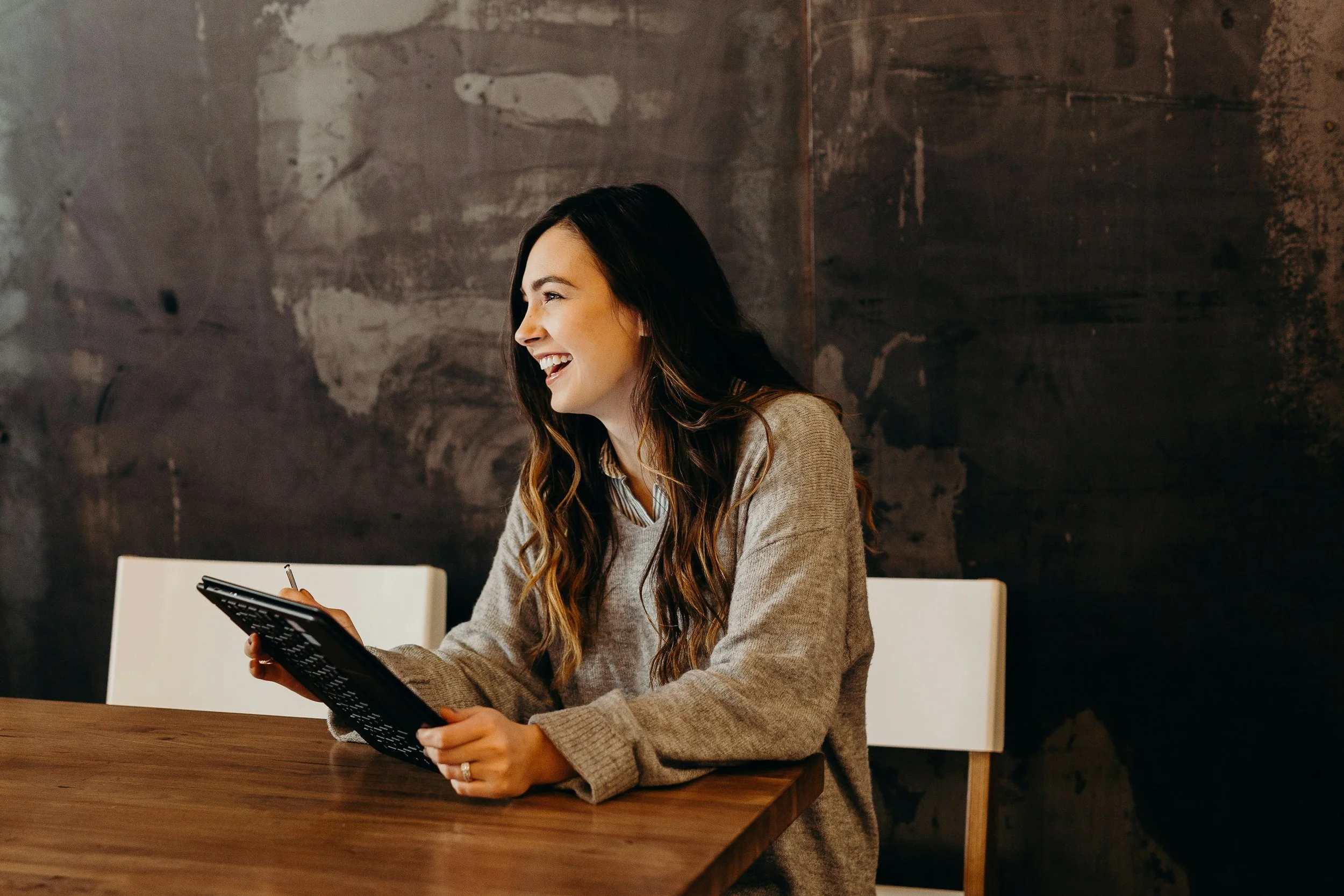 A woman with long brown hair, wearing a beige sweater, sitting at a wooden table, smiling and looking at her tablet, with a dark textured wall in the background.