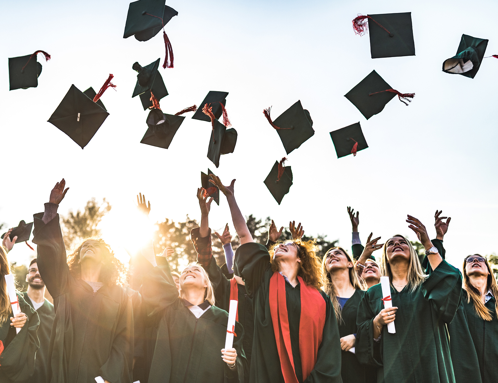 Graduates tossing their caps in the air during a graduation ceremony outdoors against a bright sky with the sun setting.