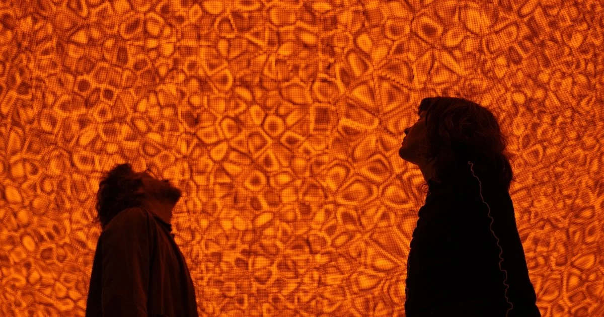Two people facing each other with an intricate orange patterned background.