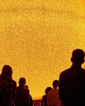 Several people in silhouette viewing a large, illuminated round display or ceiling with intricate patterns and warm yellow-orange lighting.