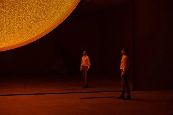 Two people standing in a dark, spacious room with a large, illuminated spherical object above them.