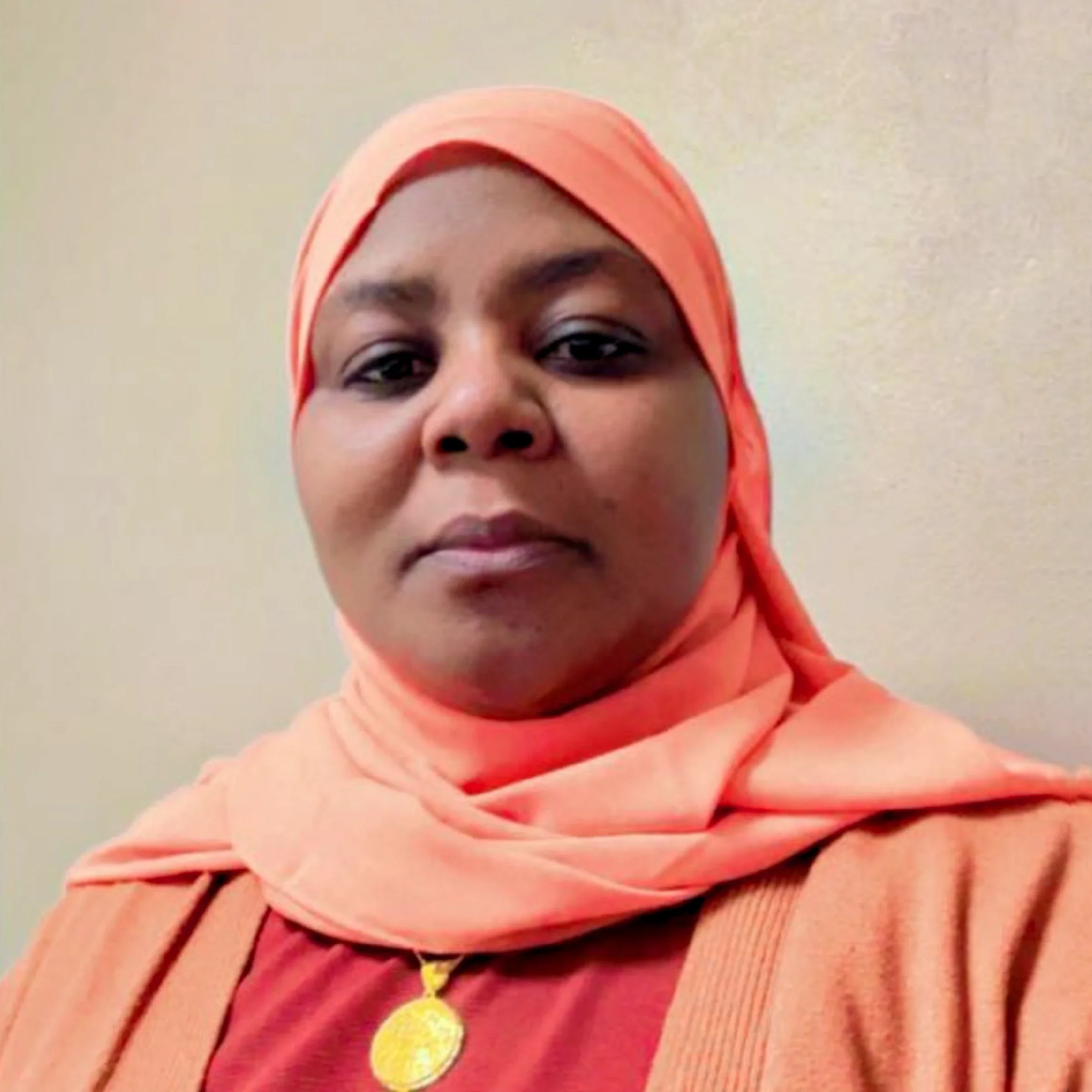 Close-up of a woman wearing a salmon-colored hijab, with a gold necklace and smiling.