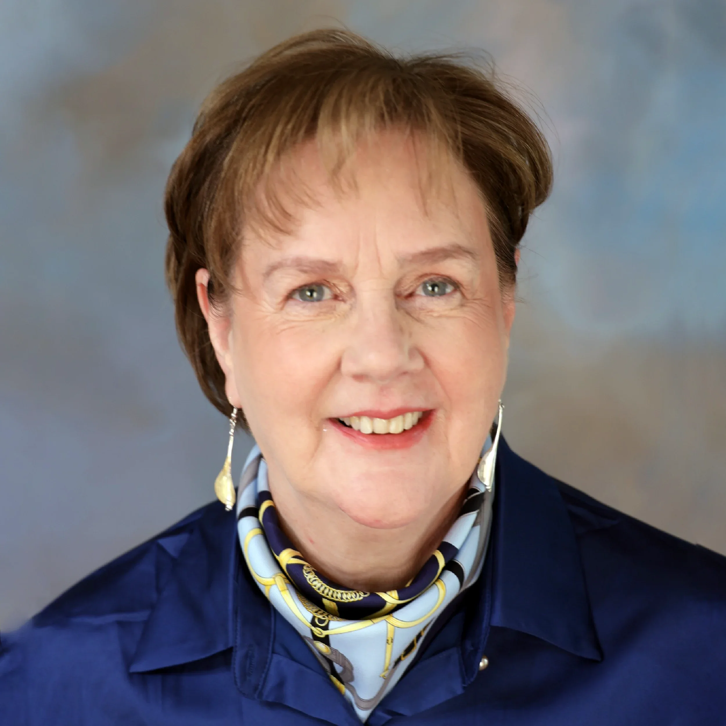 Close-up of a person's face with short brown hair, wearing a dark blue jacket and a scarf with yellow, white, and dark blue stripes, smiling at the camera against a plain gray background.