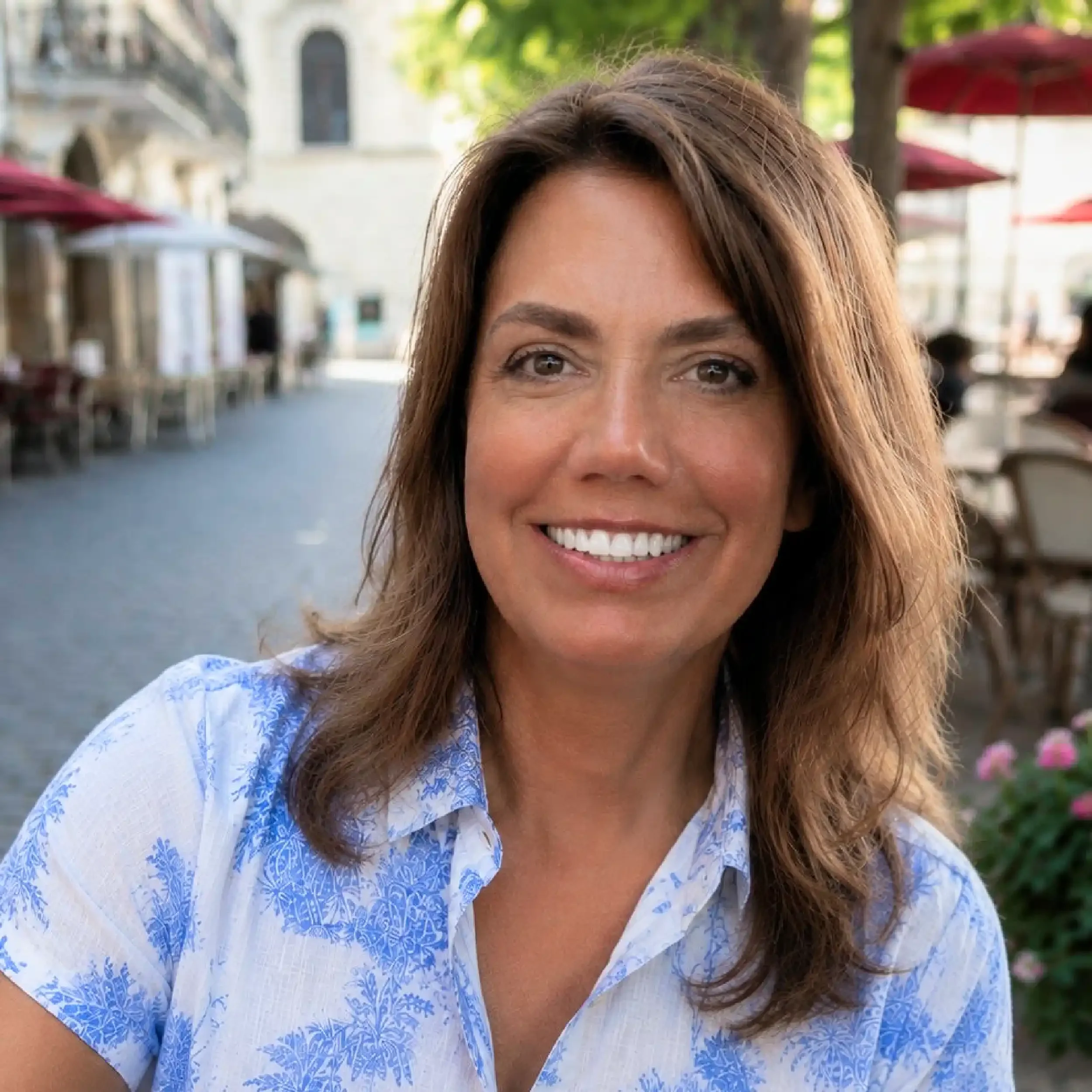 Close-up of a woman with brown hair and brown eyes smiling outdoors, wearing a blue and white patterned blouse with outdoor seating and umbrellas in the background.