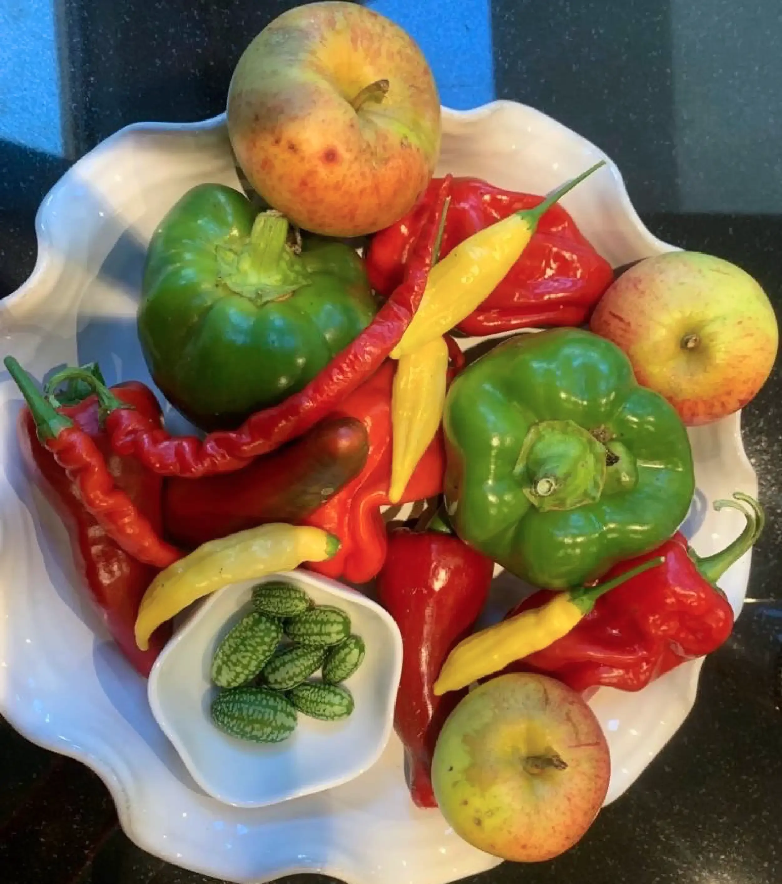 A white decorative dish filled with apples, bell peppers, peppers, and watermelon slices, placed on a dark surface.