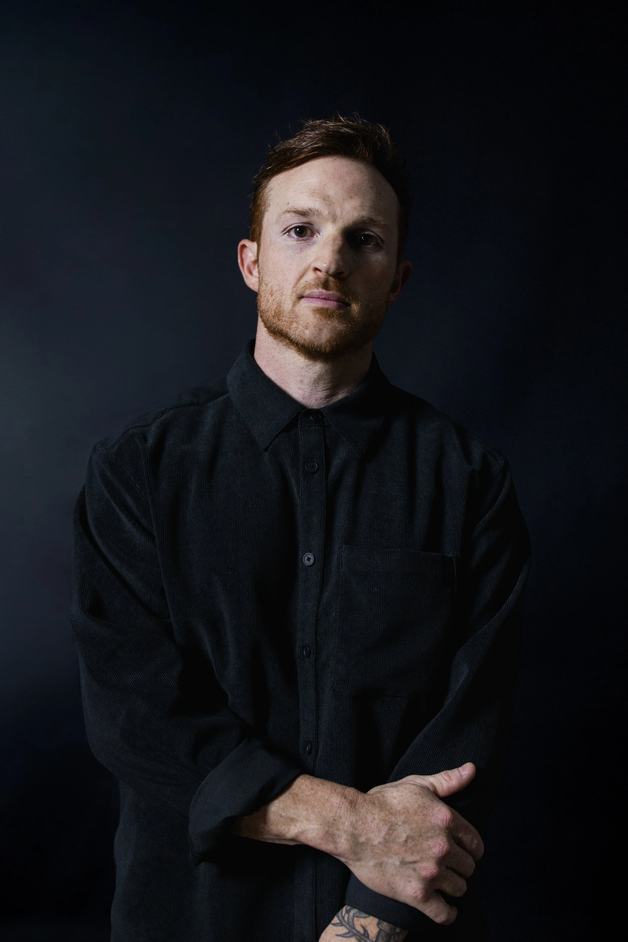 Portrait of a young man with red hair and a beard, wearing a black shirt, standing against a dark background.