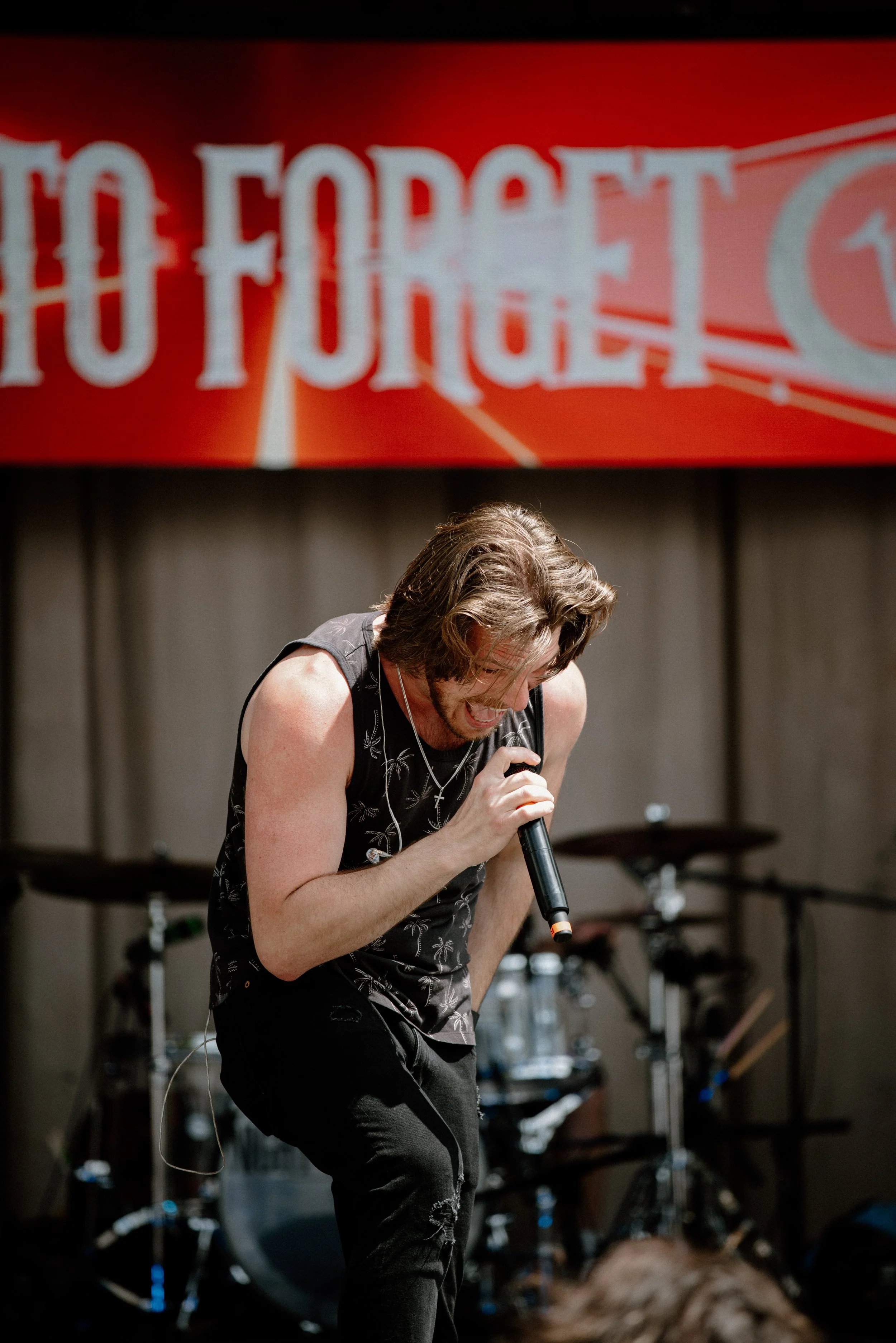 Man performing on stage, holding microphone, with drums in background, red banner above with partially visible text.