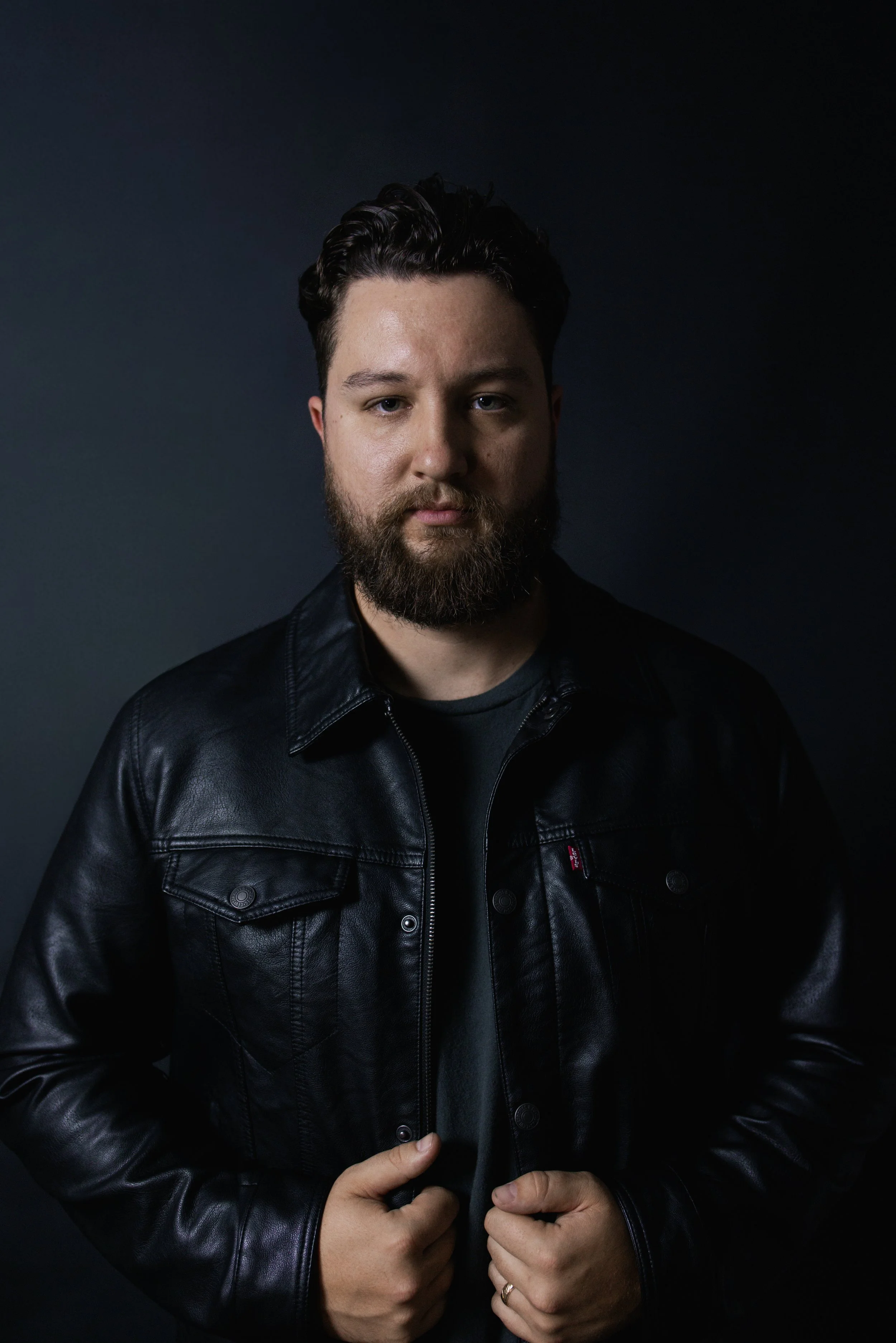 Portrait of a man with dark hair and beard wearing a black leather jacket against a dark background.