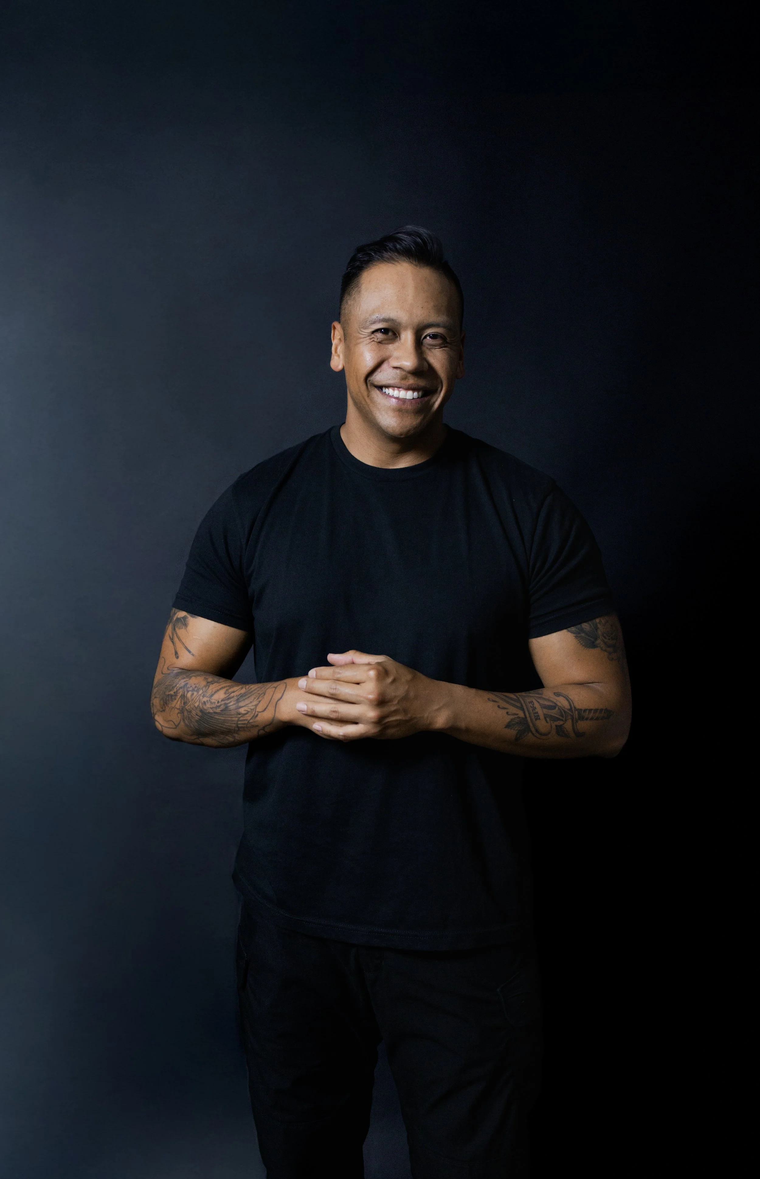A smiling man with tattoos on his arms, wearing a black t-shirt and black pants, standing against a dark background.