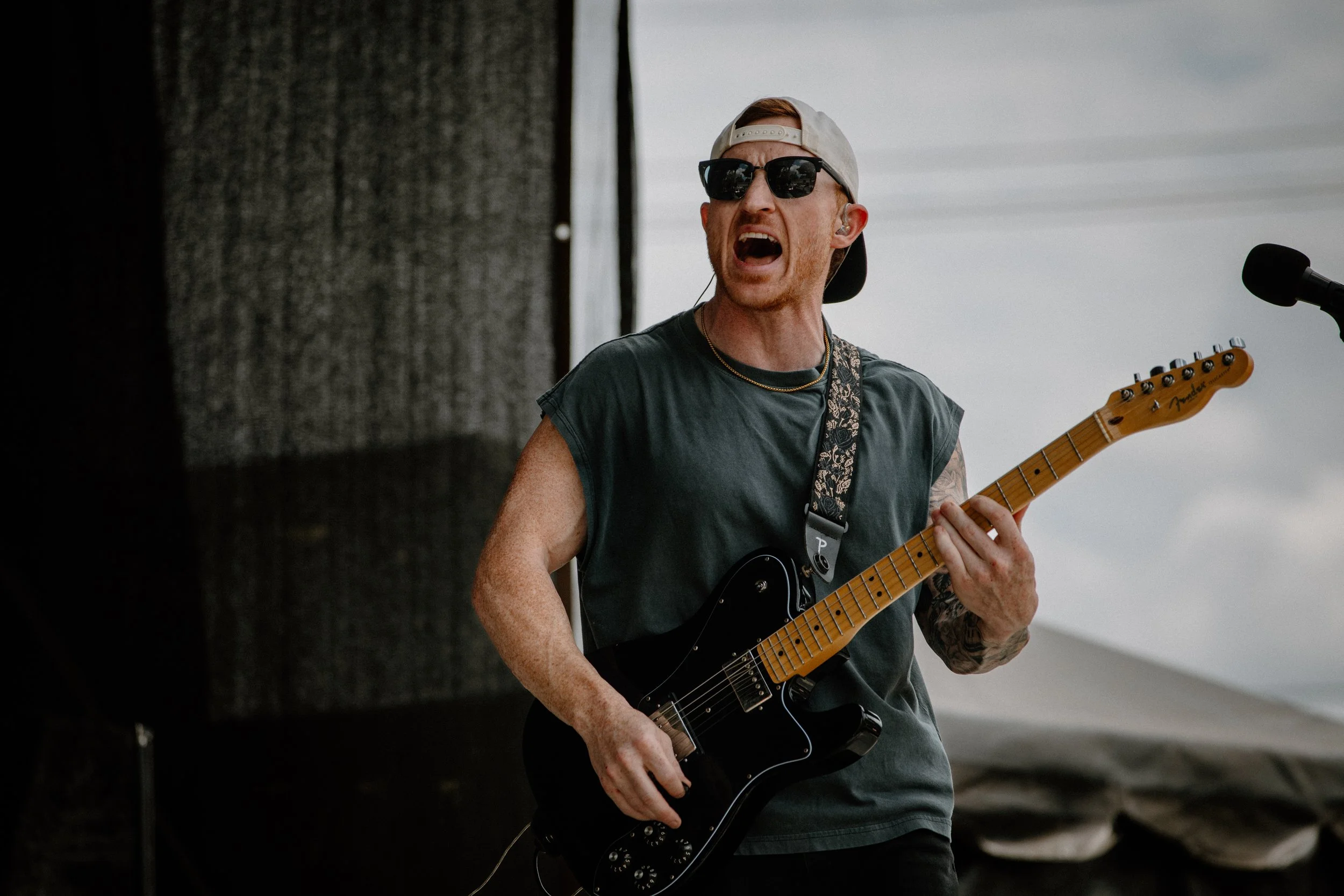 A man wearing sunglasses, a hat backwards, and a sleeveless shirt is playing an electric guitar on an outdoor stage, with a microphone nearby and a cloudy sky in the background.