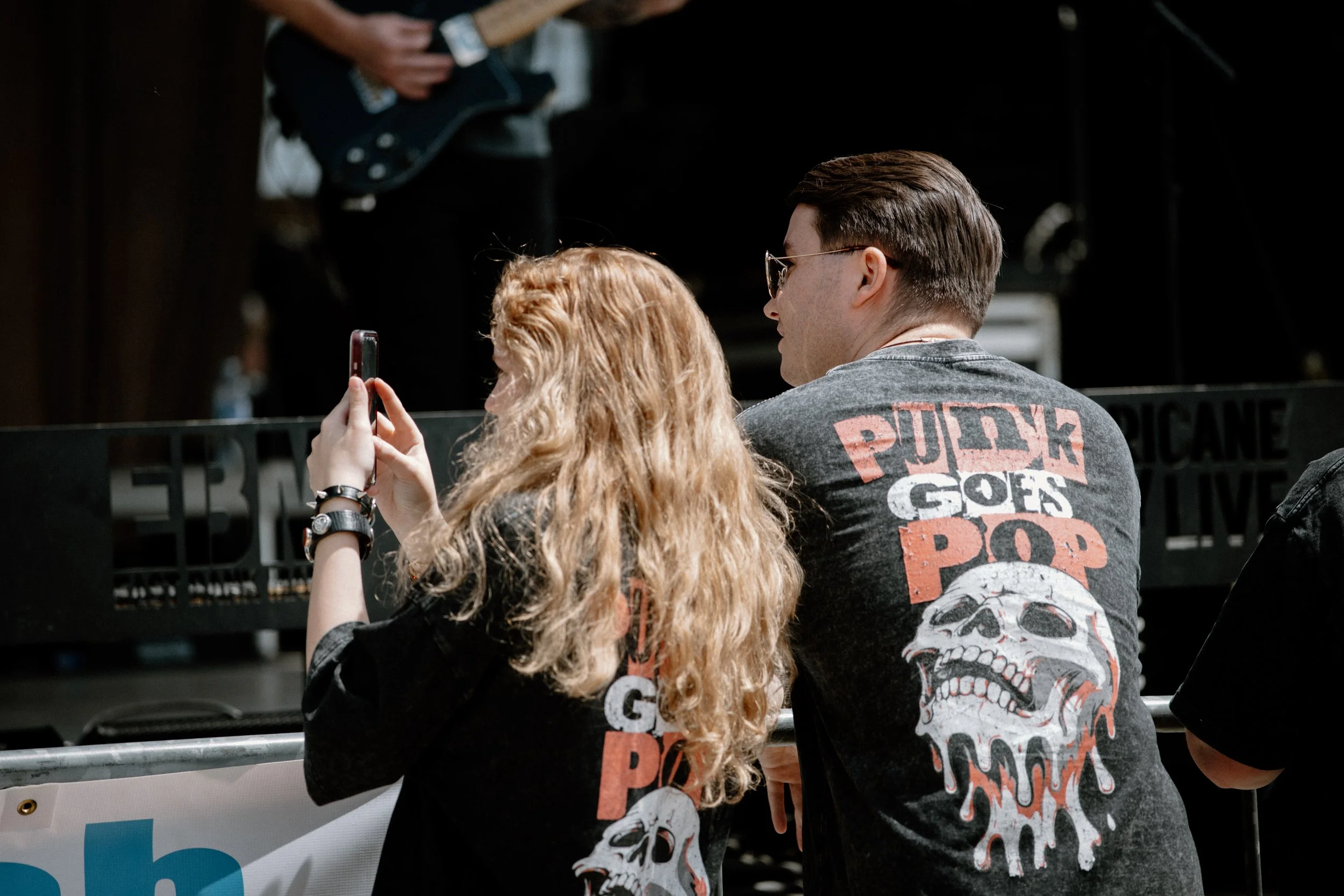 People at an event, one person with long curly red hair taking a photo with a smartphone, another person with short dark hair wearing sunglasses and a black T-shirt with a skull graphic and text. There is a person playing an electric guitar in the ba