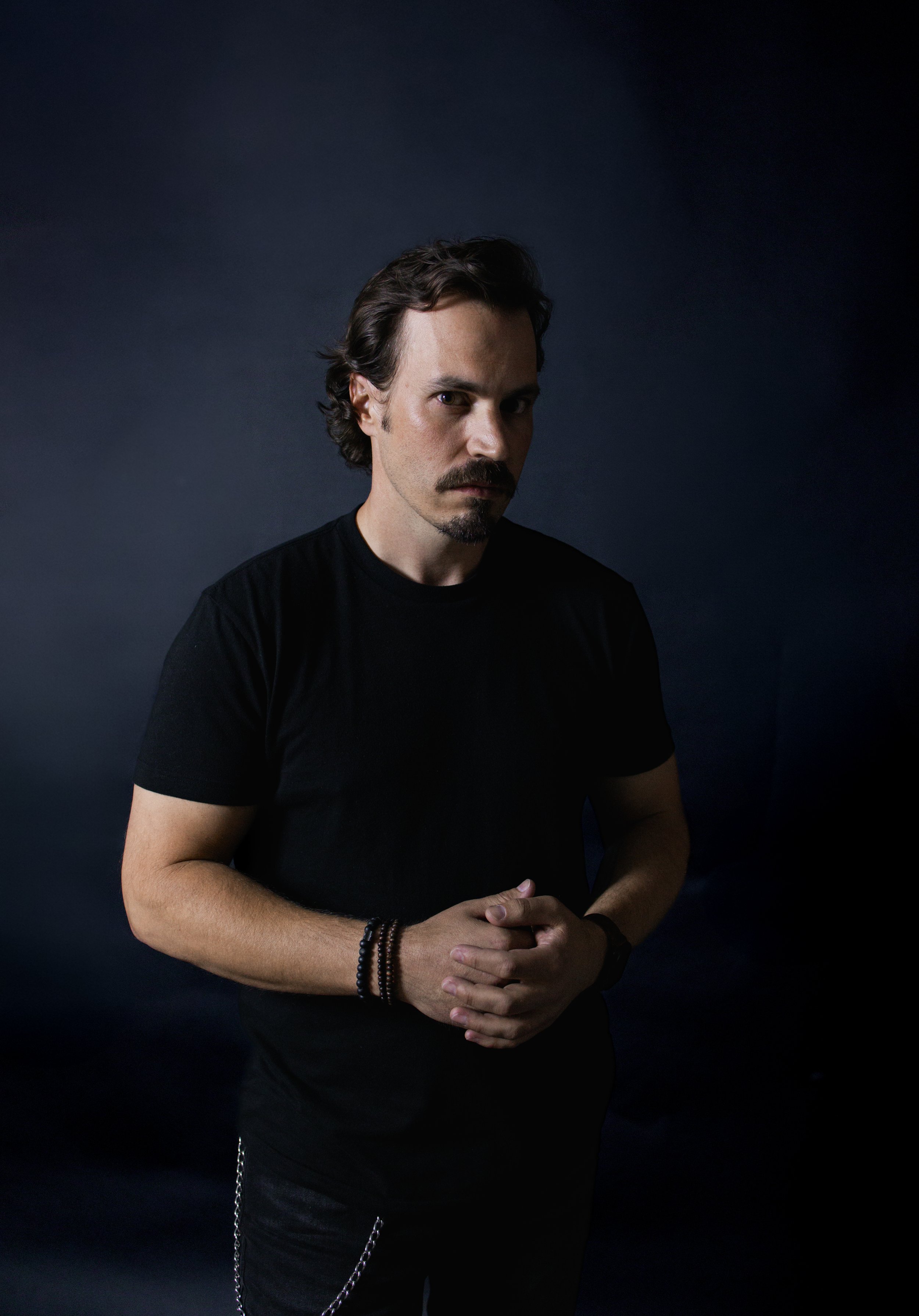 A man with dark, wavy hair and facial hair wearing a black t-shirt, with his hands clasped in front of him against a dark background.