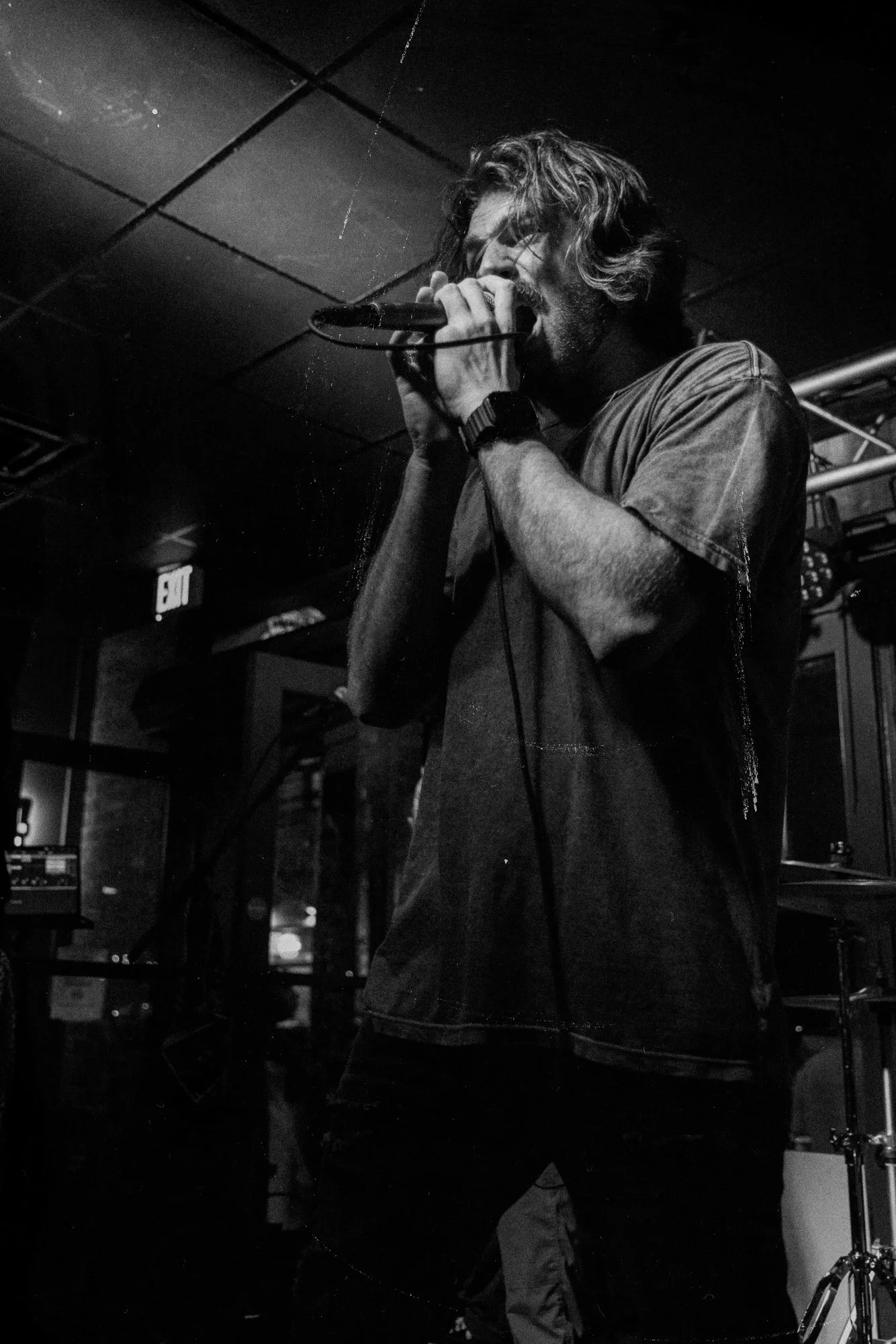 A man with long hair and a beard singing into a microphone in a music venue with low lighting and musical equipment.