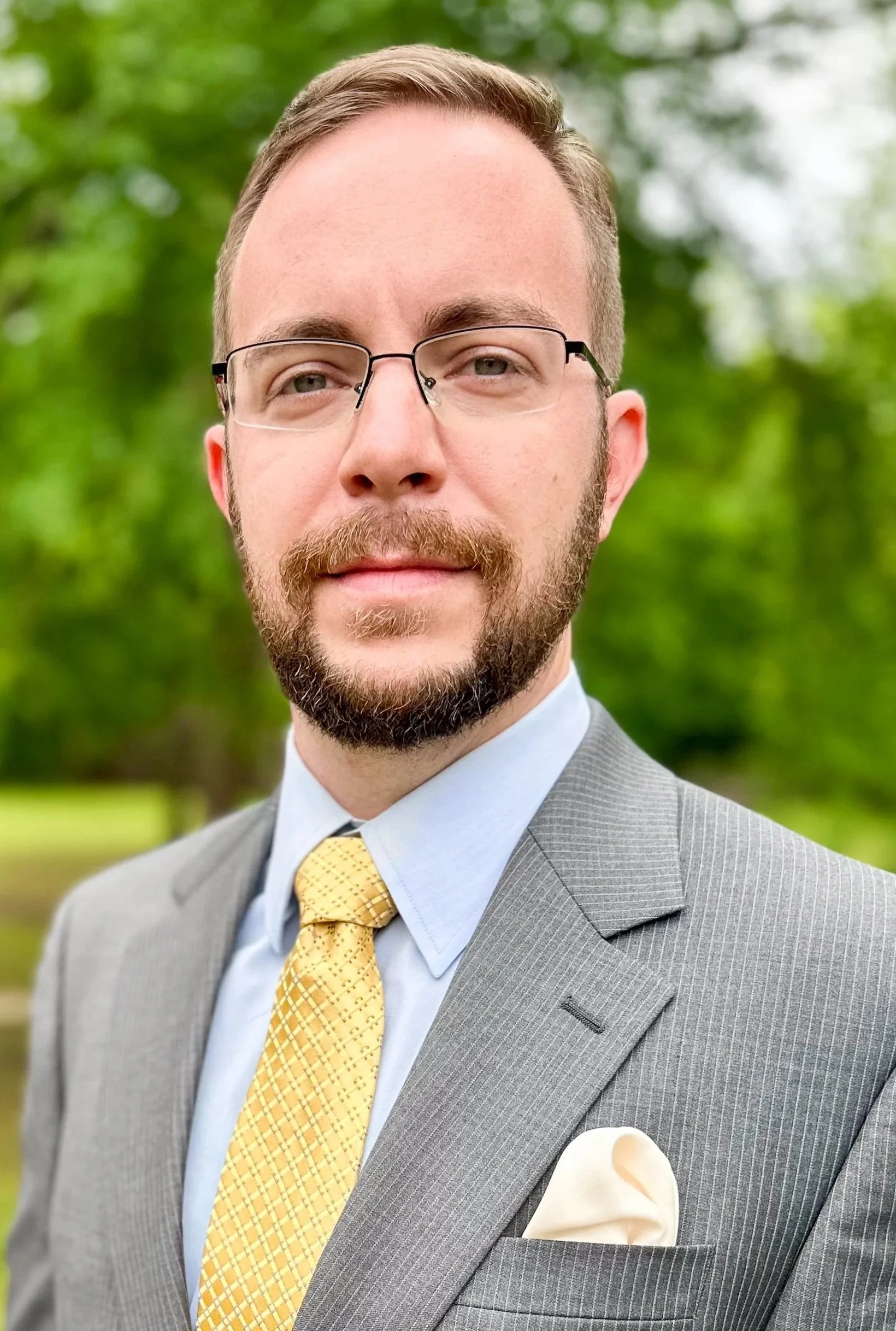 A man wearing glasses, a gray suit, light blue shirt, and yellow patterned tie, standing outdoors with a green, blurry background.